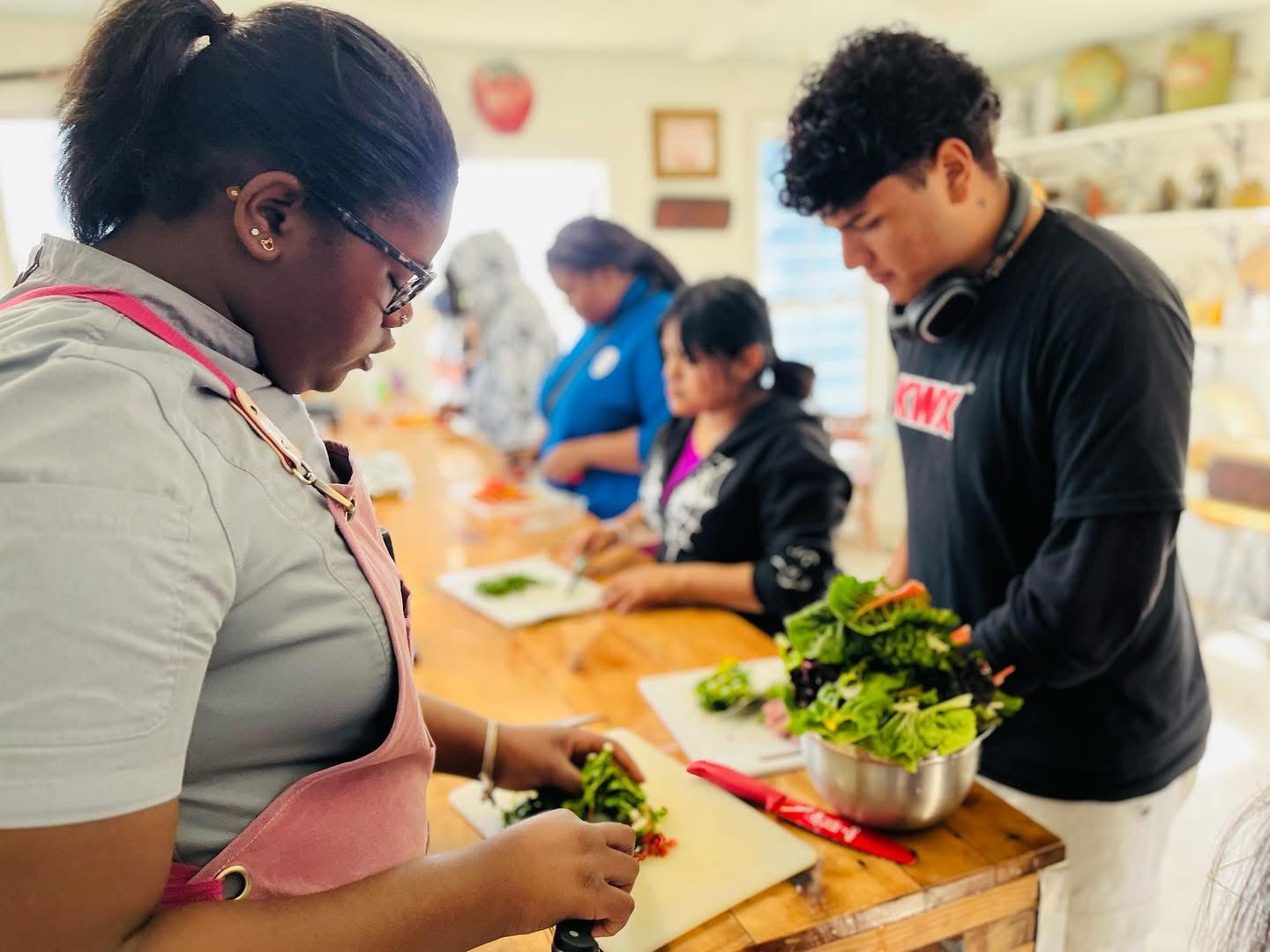 People preparing vegetables at a counter in a kitchen or cooking class, with others in the background. 20 words.