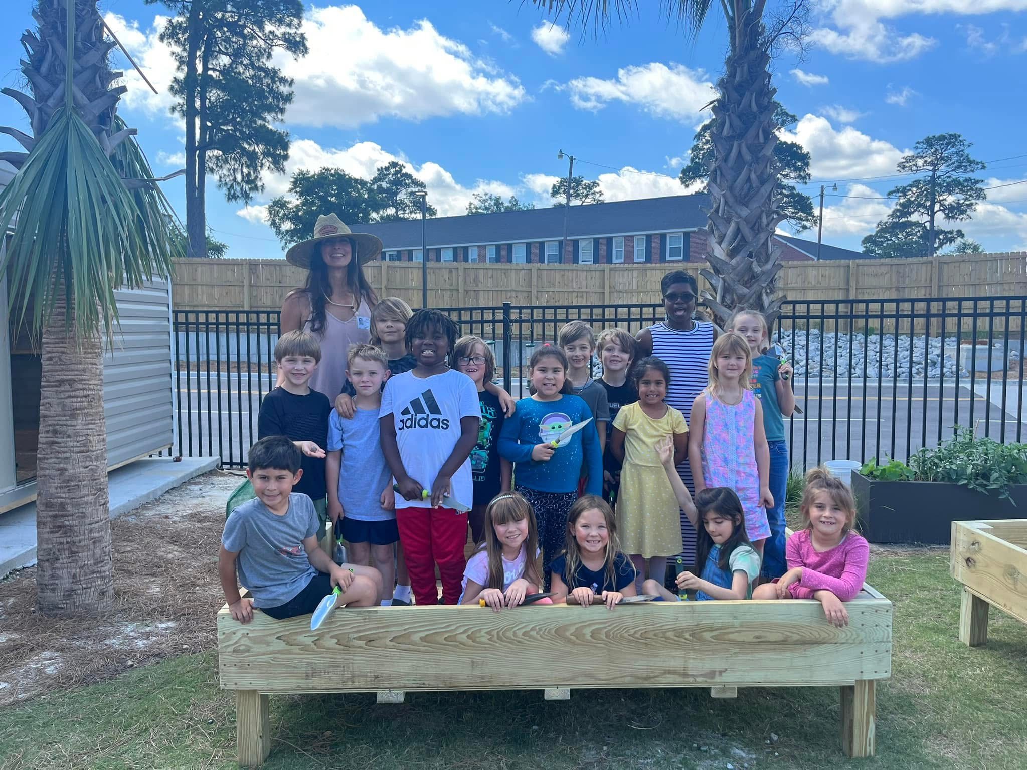 Group of children and an adult outdoors near a wooden planter box, with trees and a building in the background.