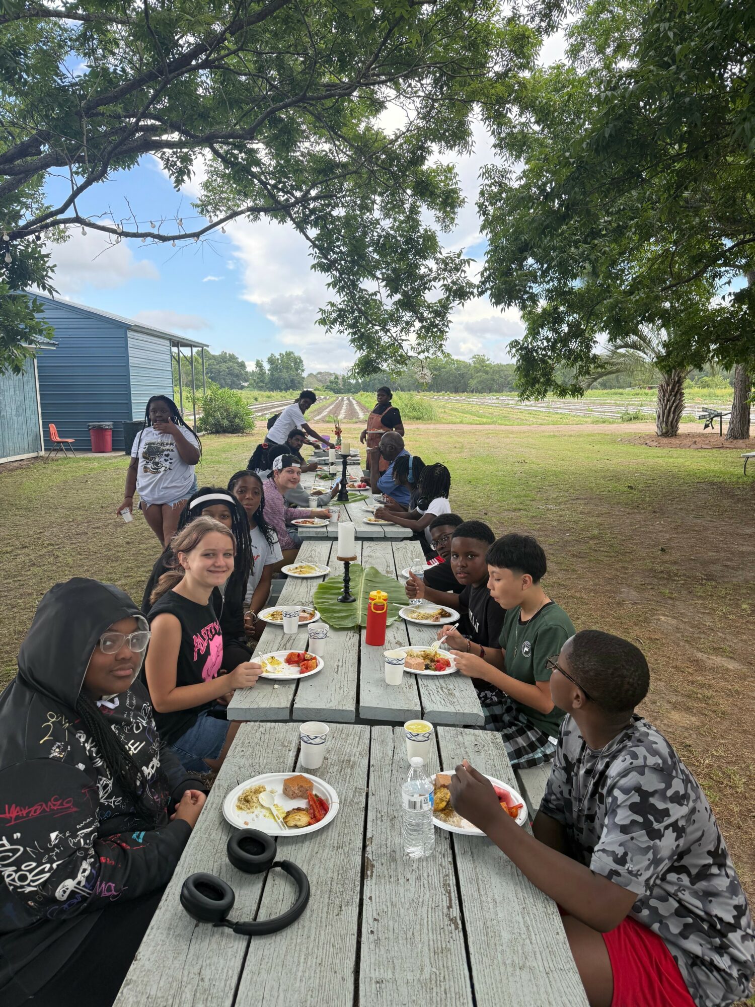 People sitting at a long outdoor picnic table under trees, eating food, with a blue building in the background.