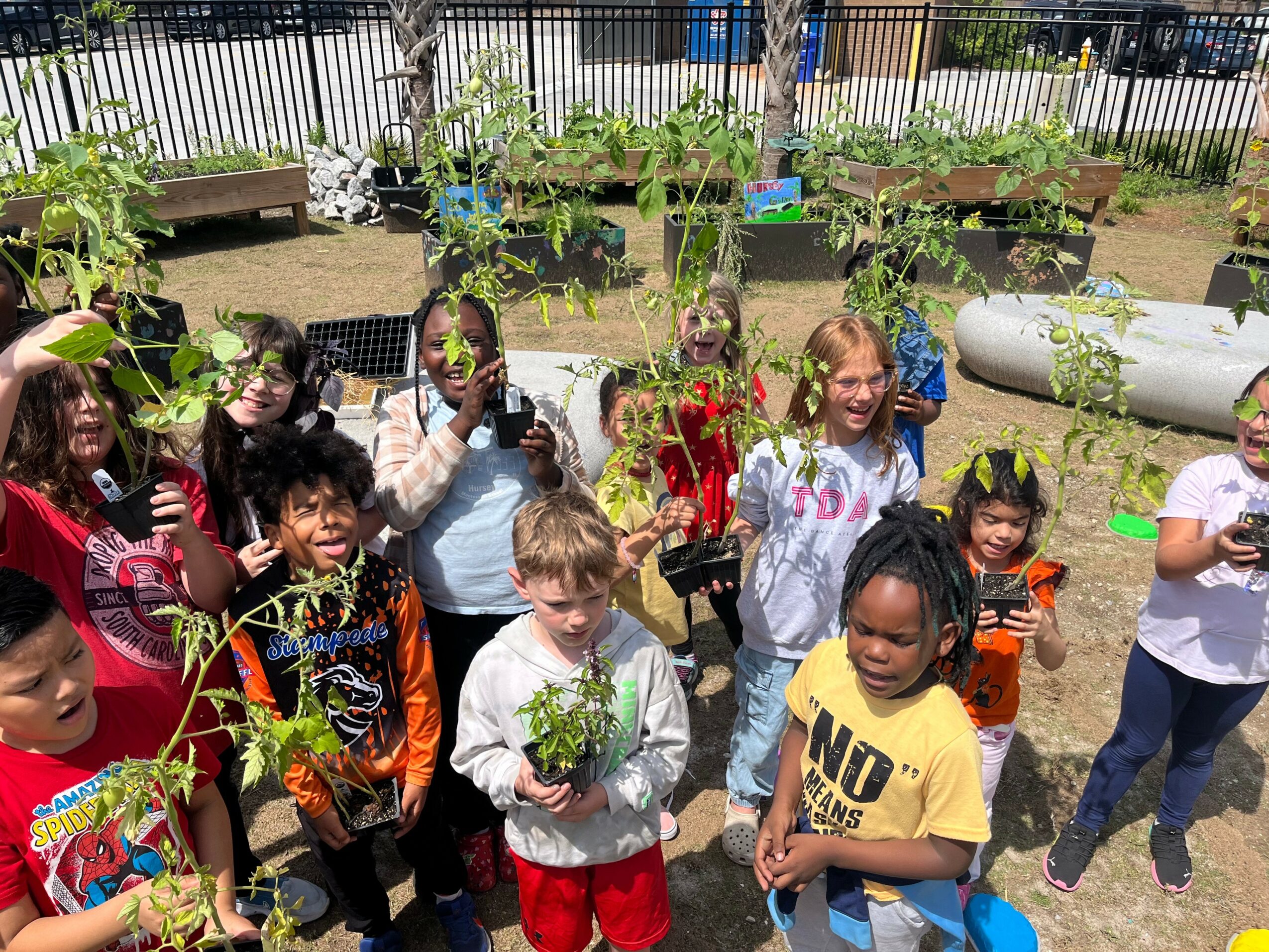 Group of children outdoors in a garden, holding plants and smiling, with a fence and garden beds in background.
