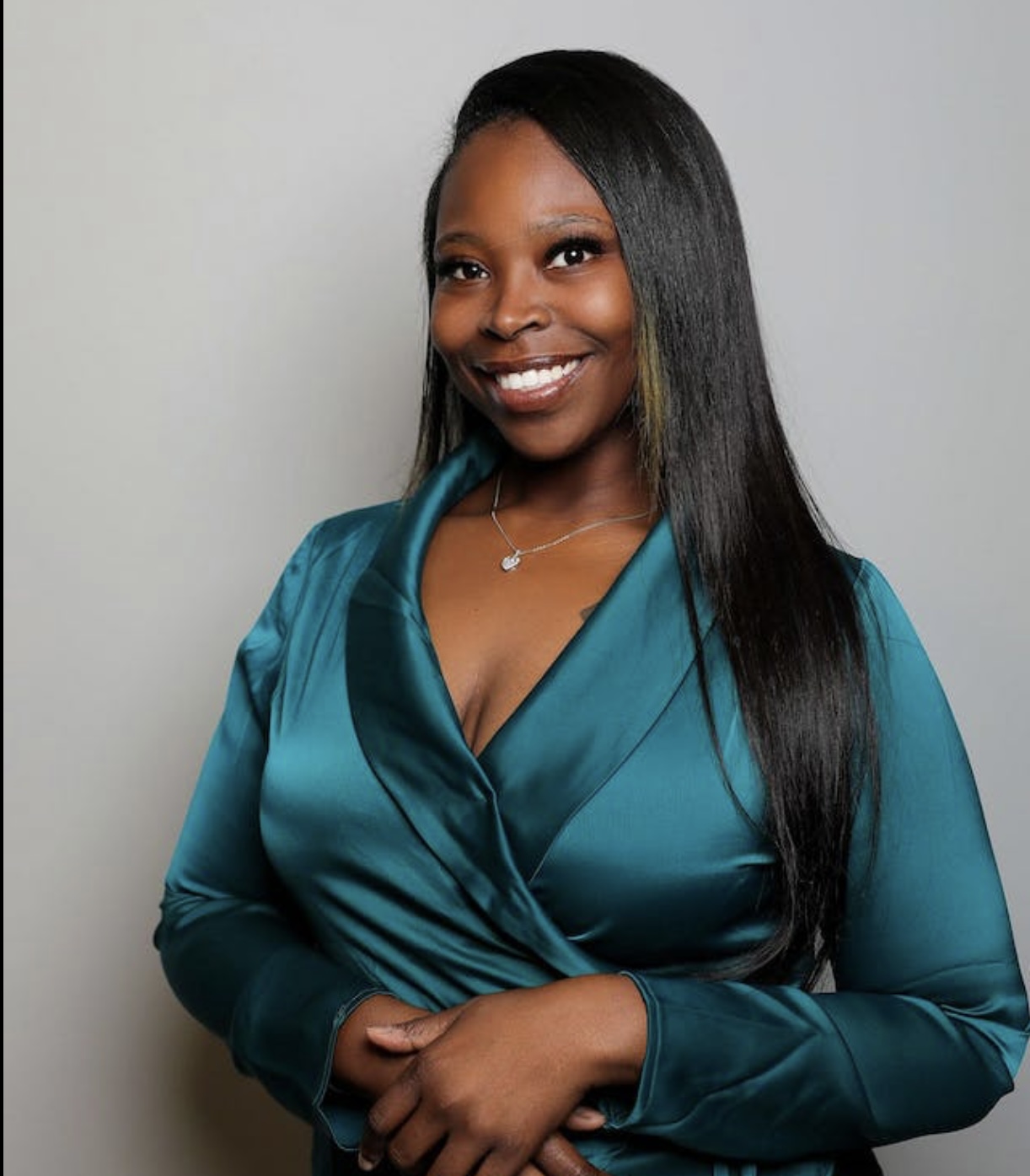 Smiling woman with long black hair wearing a teal satin blouse, standing against a light gray background.