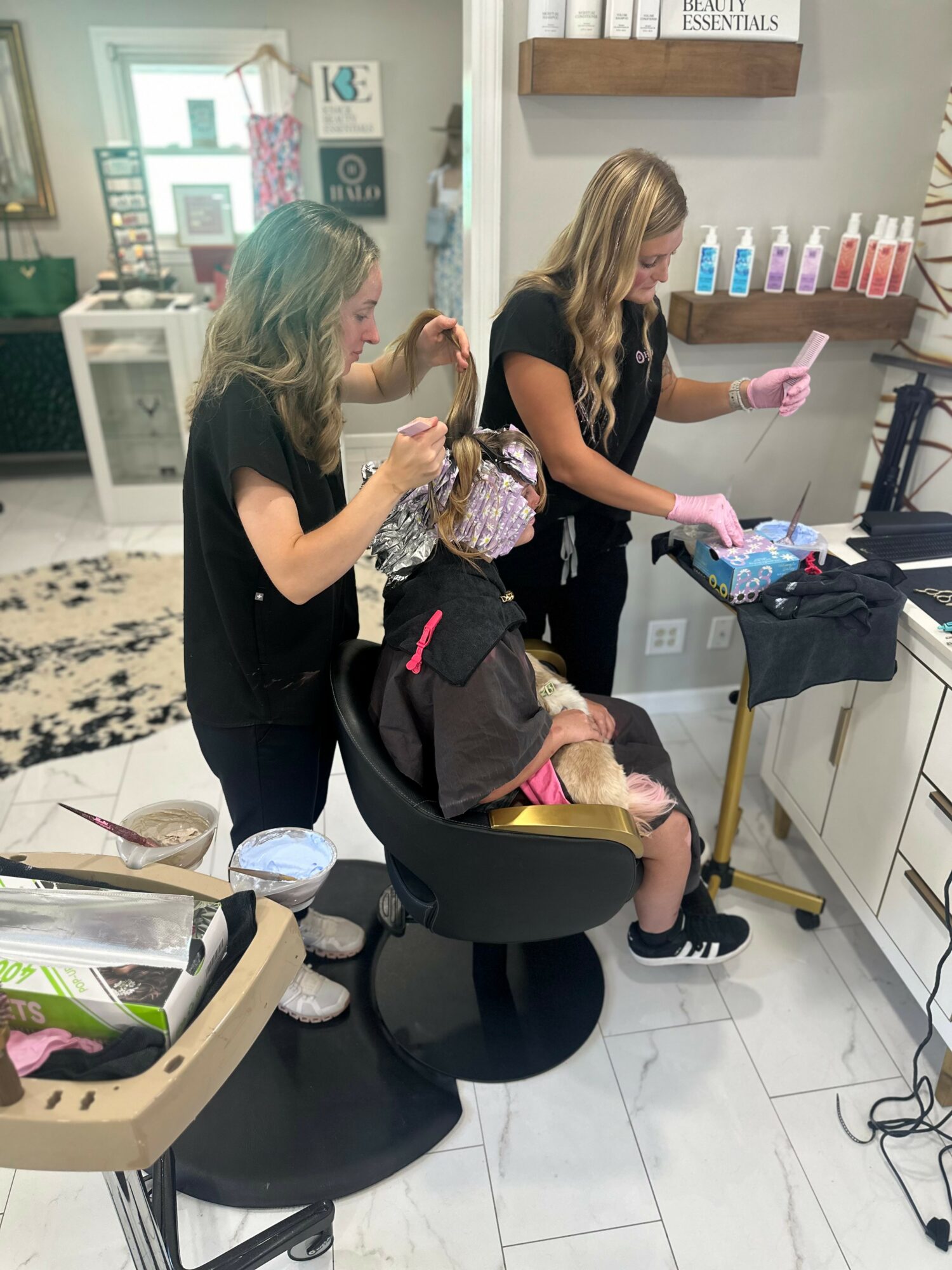 Two women working on a child's hair in a salon, with hair products on the wall and salon tools nearby.