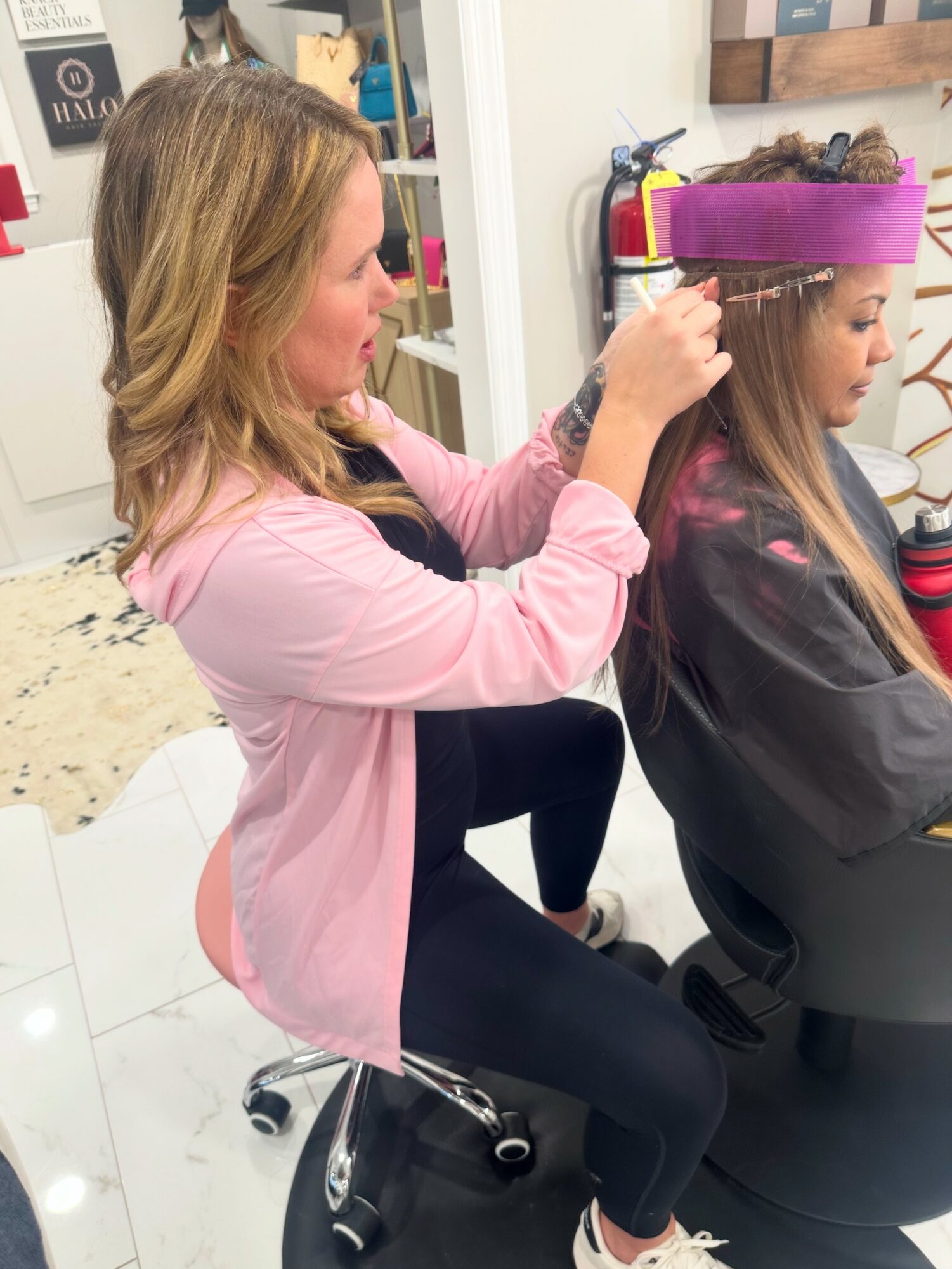 Woman styling a young girl's hair in a salon, girl wearing a purple headband, woman in pink jacket, salon setting.