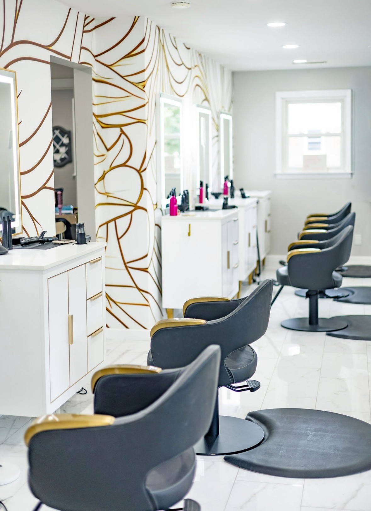 Salon with styling chairs, mirrors, and white cabinets, decorated with gold and white patterned wall, bright window light, and clean floor.