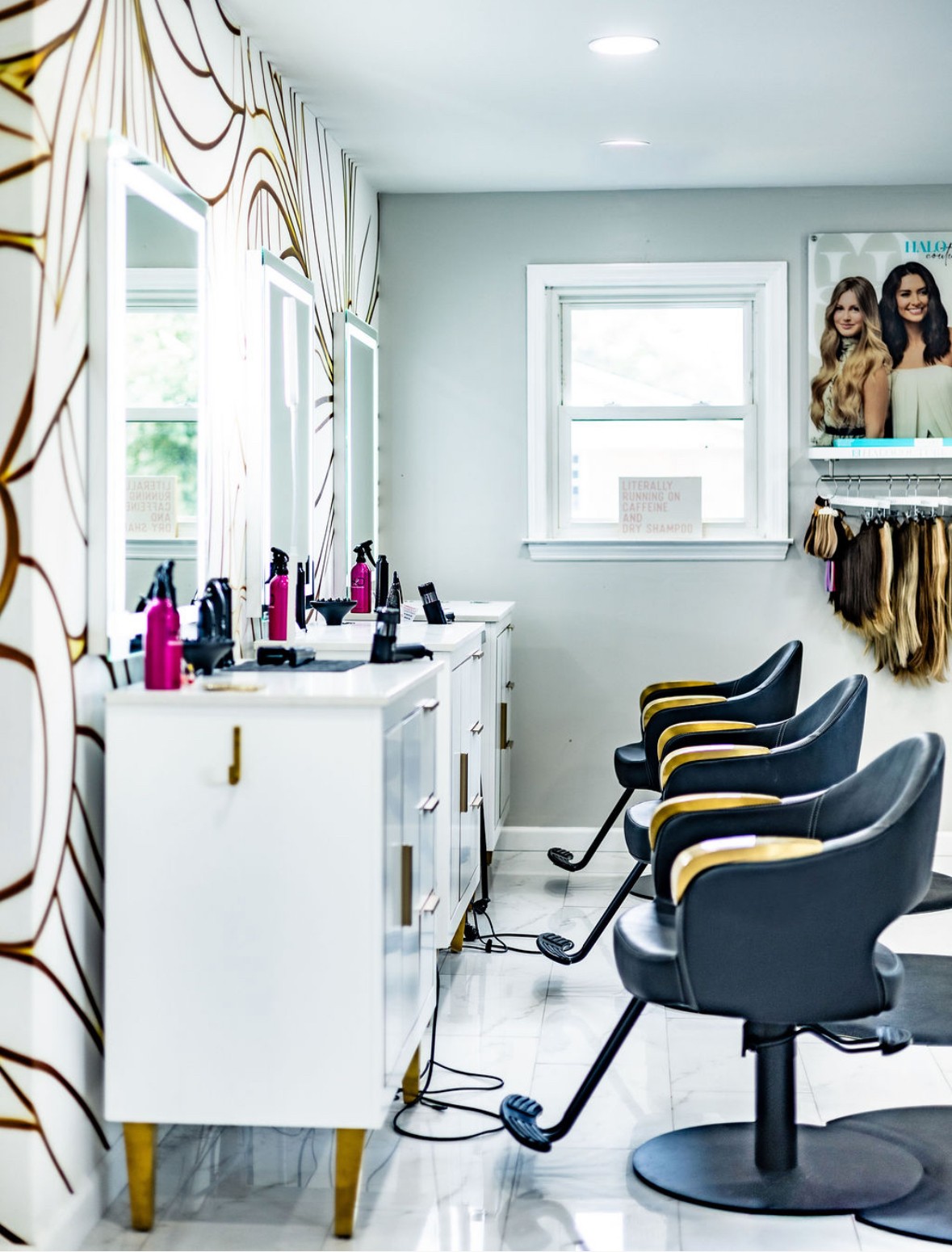 Salon interior with white cabinets, styling products, and three black chairs facing mirrors, window, and a poster on the wall.