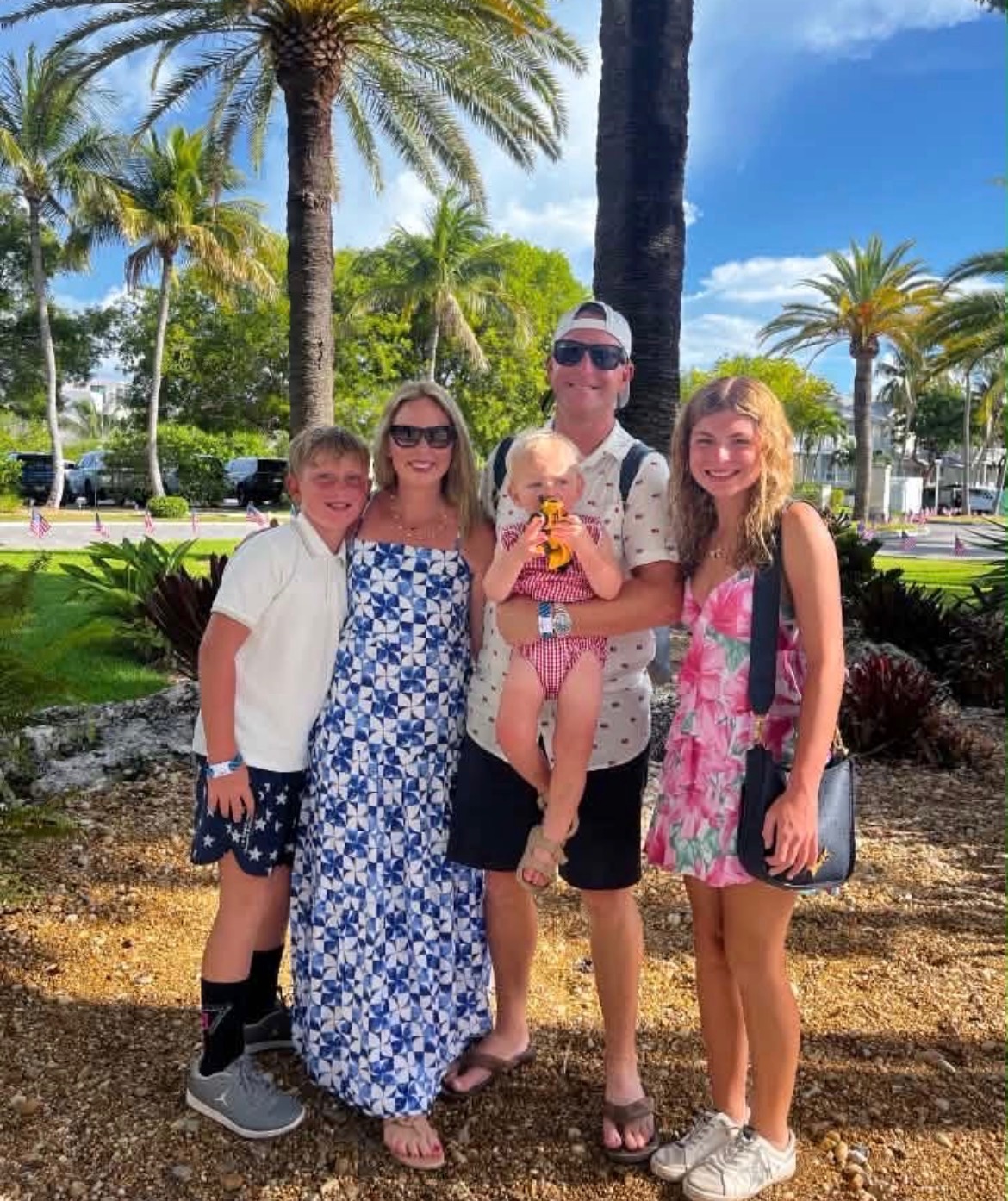 Group of five people standing outdoors with palm trees and blue sky, smiling, holding a child, in casual summer clothing.