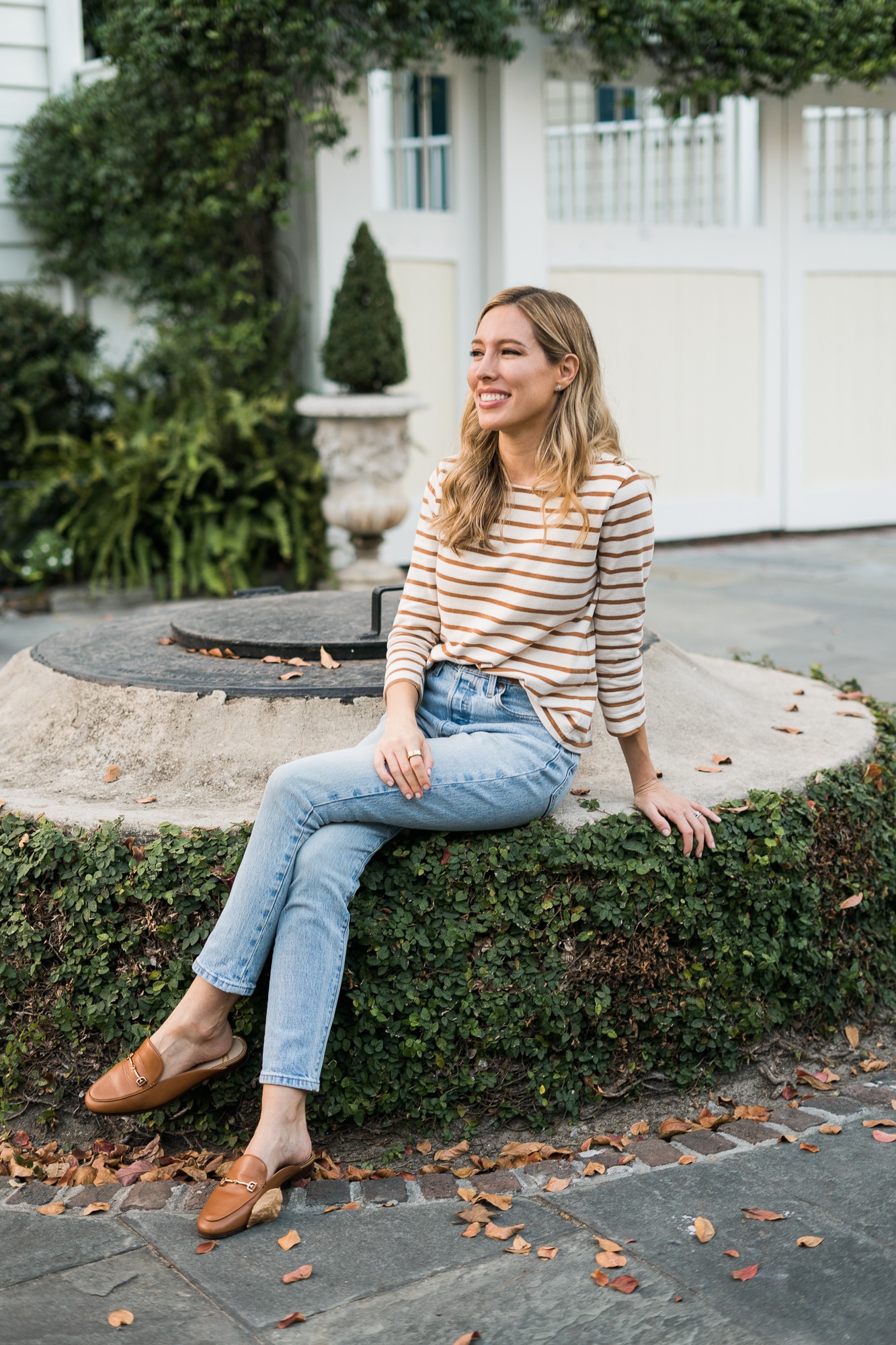 Woman sitting on a stone ledge outdoors, smiling, wearing a striped shirt and jeans, with greenery and a white house in background.