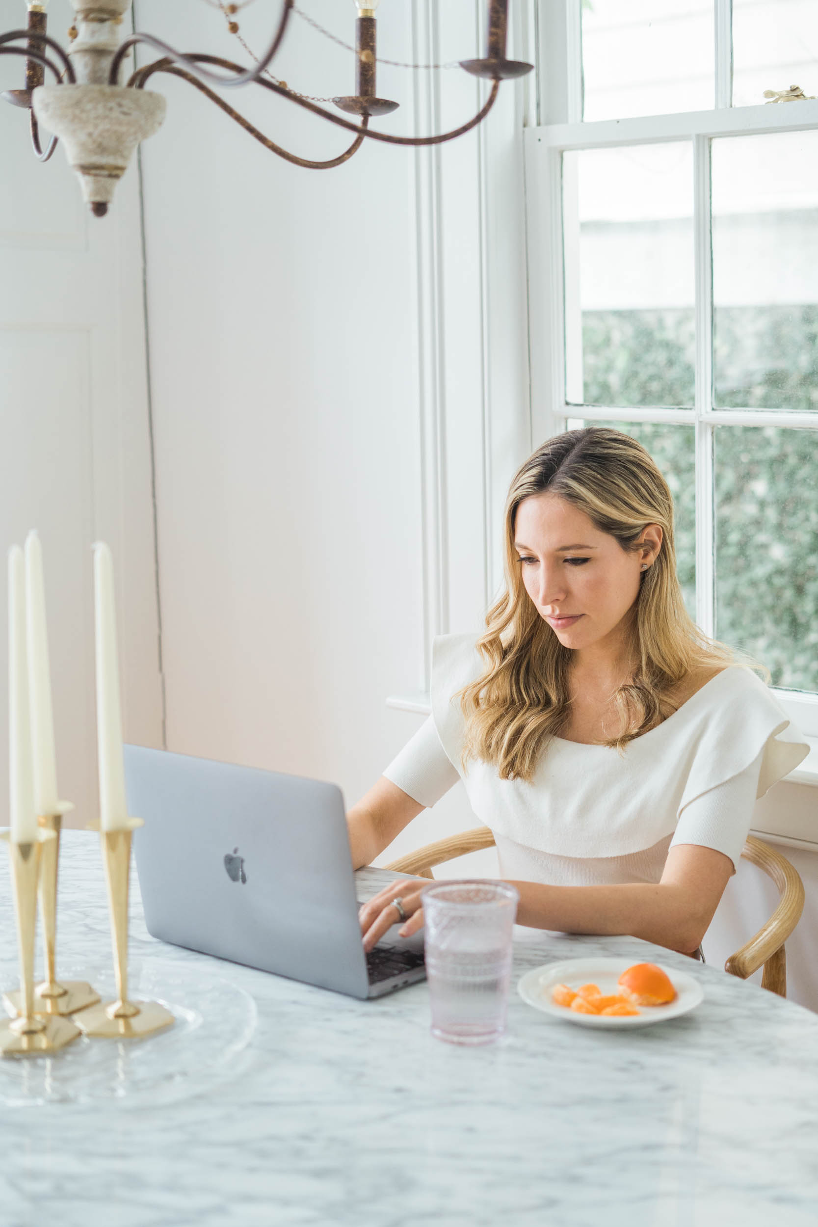 Woman with long hair working on a laptop at a marble table, with a window and greenery outside, candles on the table.