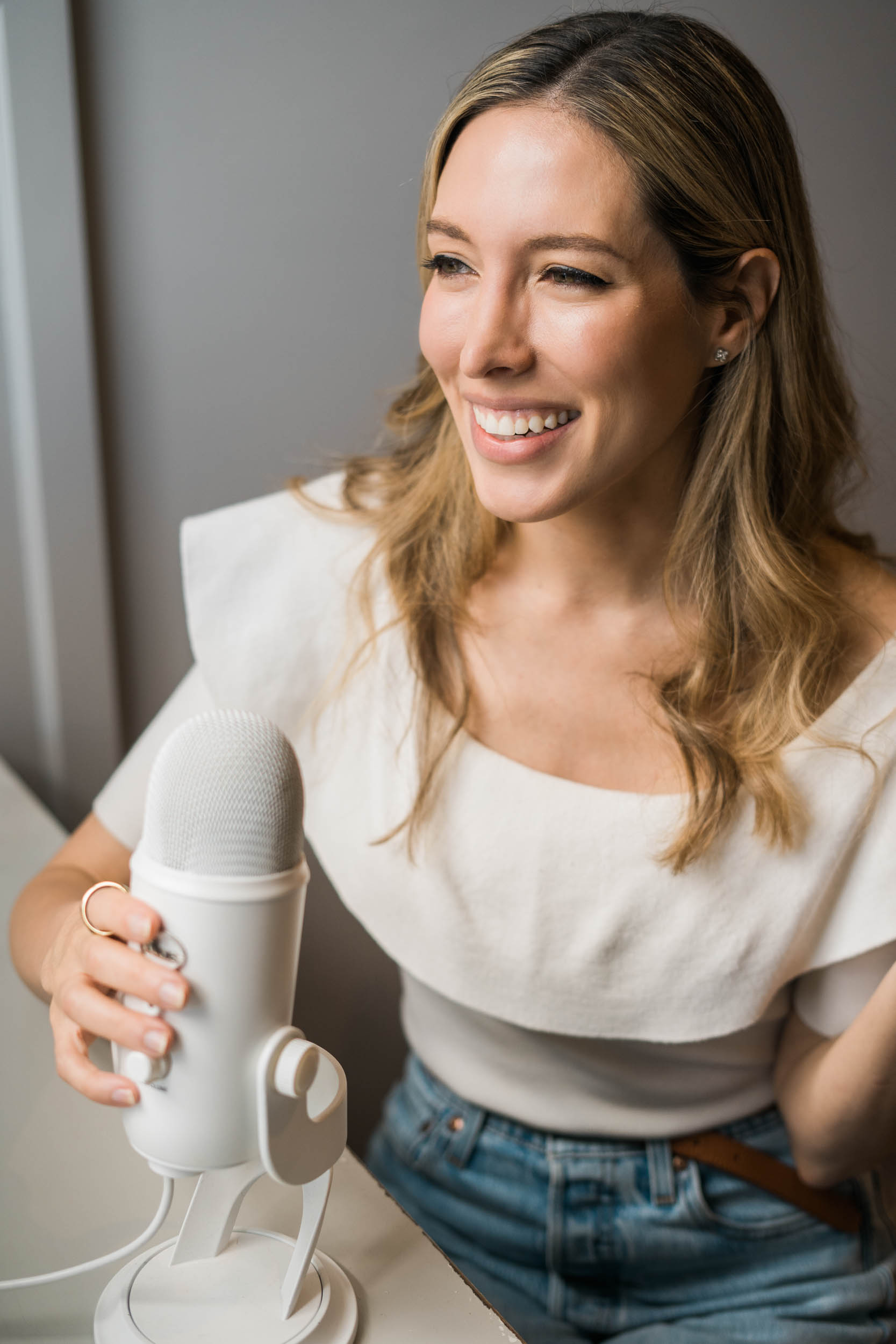 Woman smiling, holding a microphone, wearing a white top, in a well-lit indoor setting.