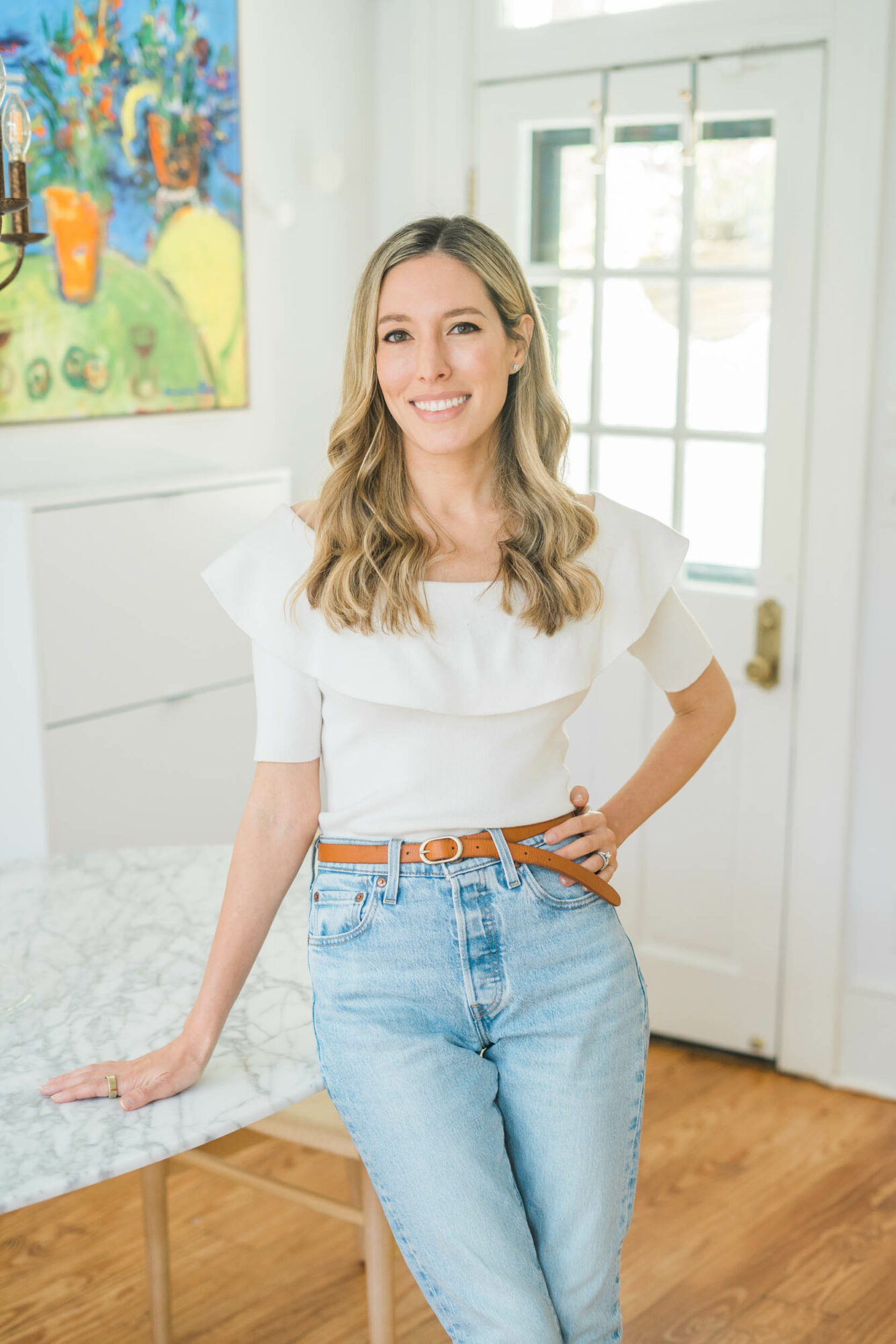 Young woman with long wavy hair smiling, standing in a bright room with a window and colorful painting, wearing a white ruffled top and jeans.