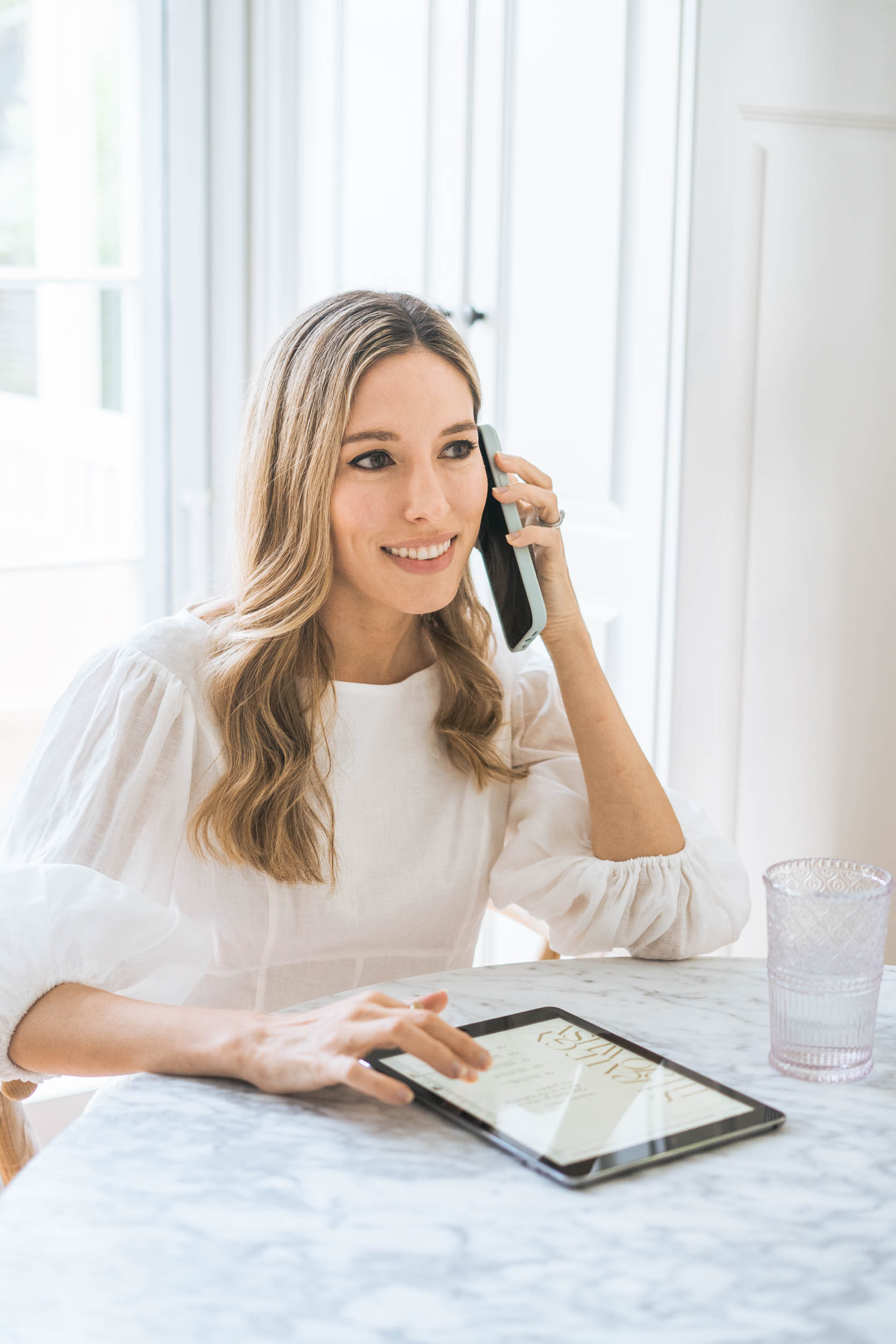 Woman with long blonde hair smiling, talking on phone, sitting at a table with tablet and glass of water.