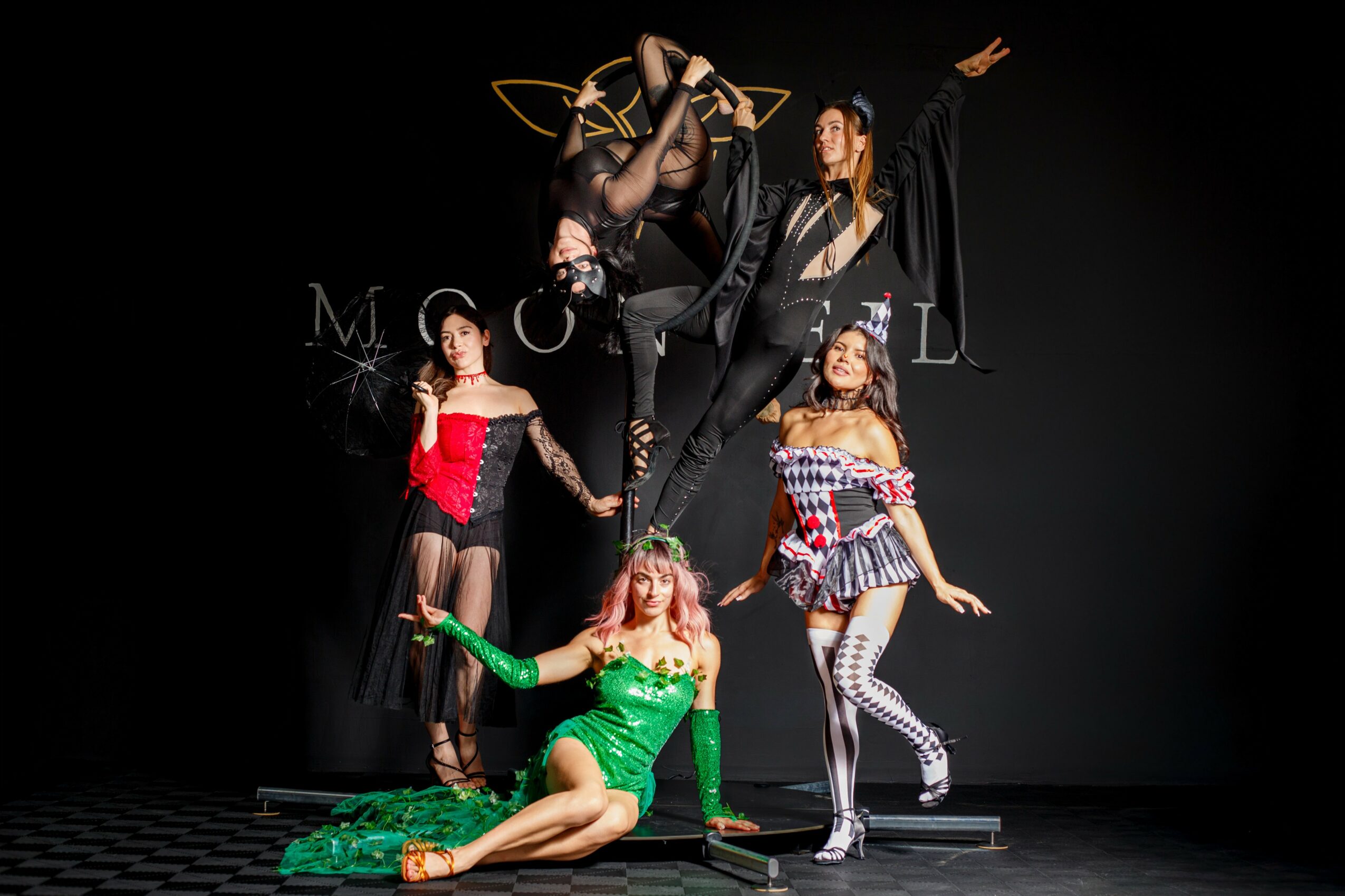 Group of five women performing on stage with a dark background, some in colorful costumes, one in green, one in red, and others in patterned outfits.