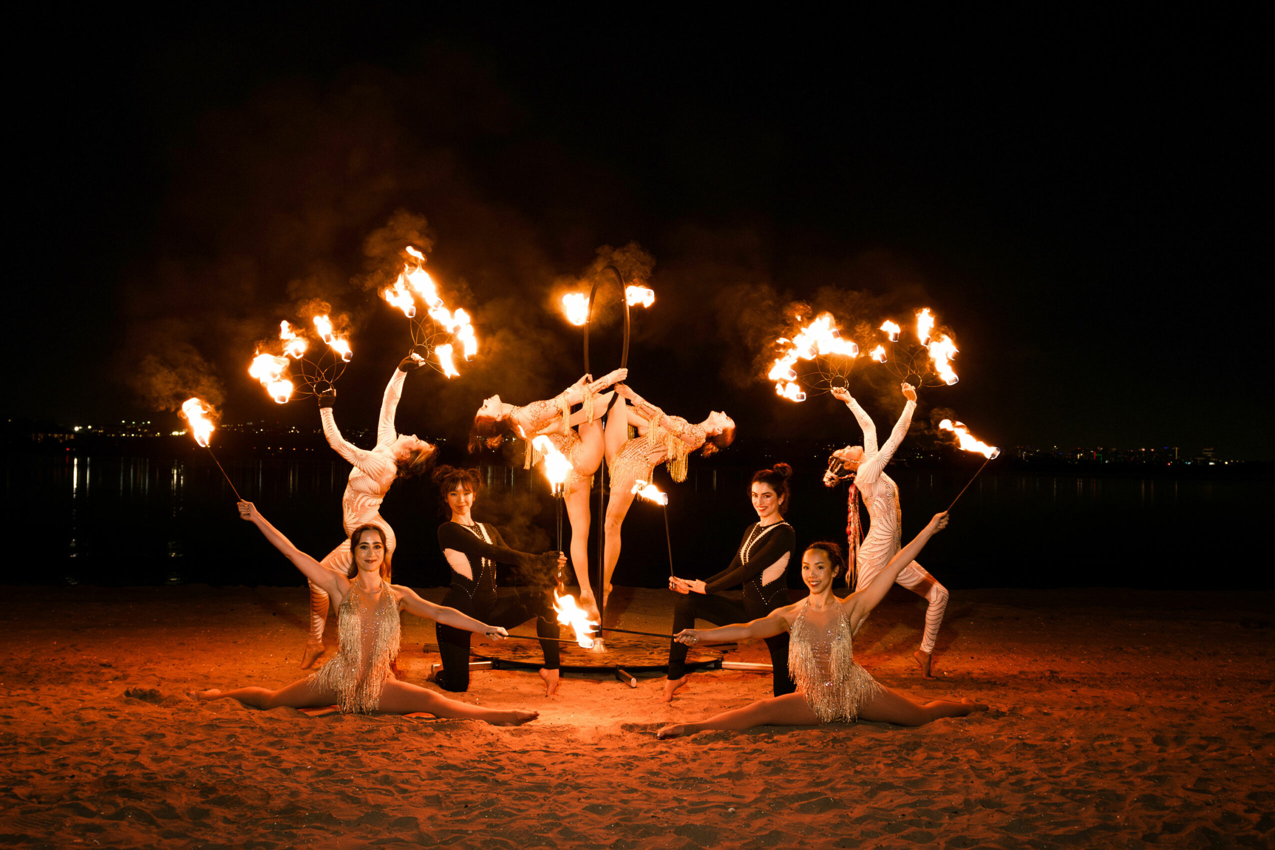 Group of performers with fire props on a beach at night, some lying and some standing, with flames in the background.