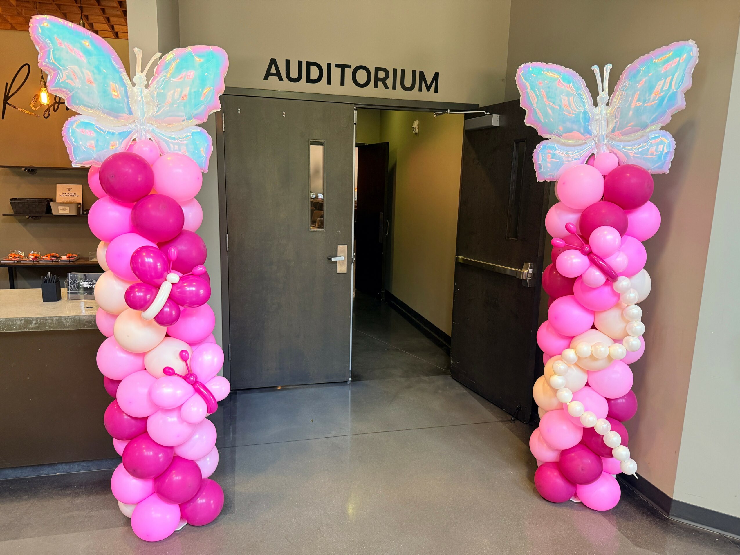 Two balloon columns with butterfly decorations at an auditorium entrance, decorated with pink, white, and purple balloons.