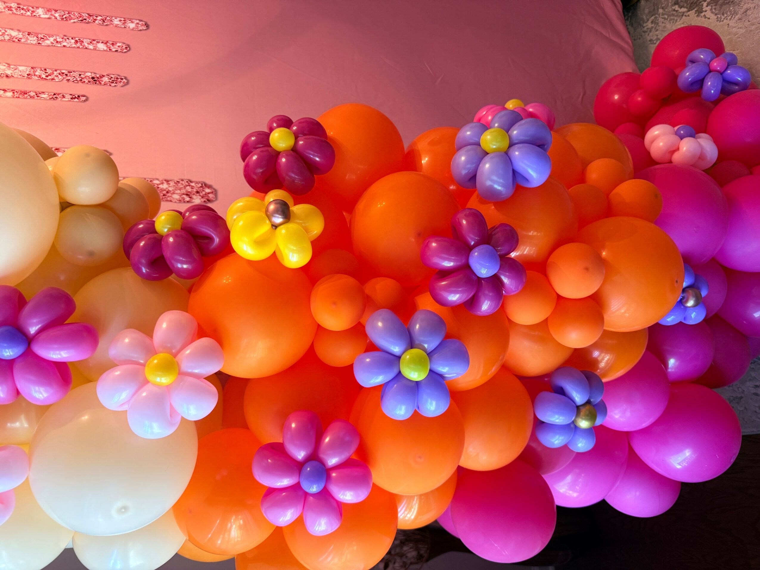Colorful balloon arrangement with orange, pink, purple, yellow, and white balloons, decorated with balloon flowers, on a pink background.