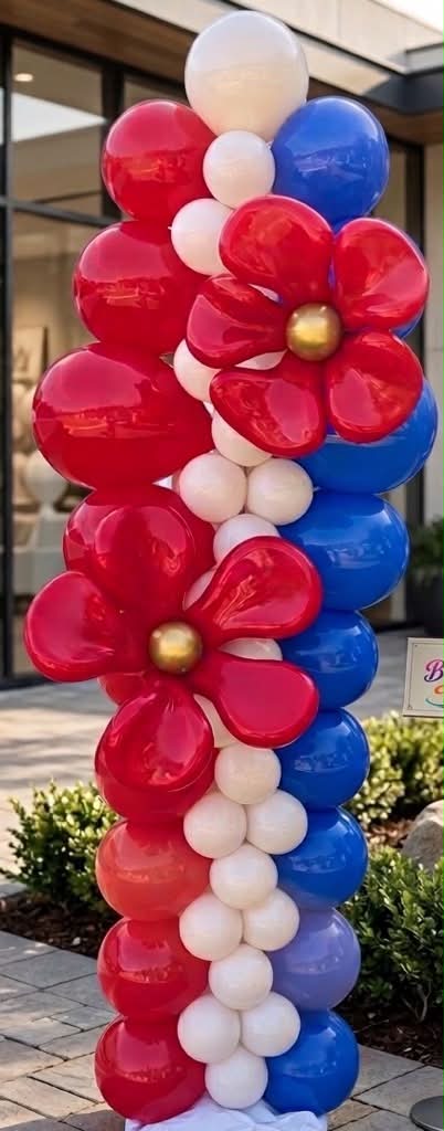 Column of red, white, blue, and gold balloons with large red flowers, outdoors near plants and a building.