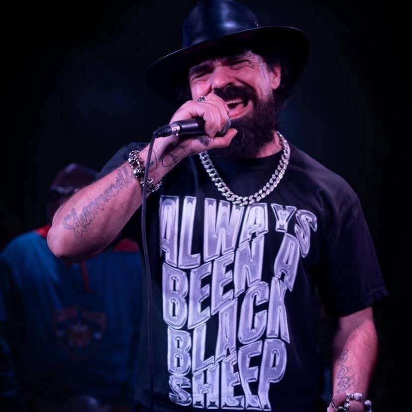 Man singing into microphone, wearing hat, chain, and a T-shirt with text, on stage with dark background.