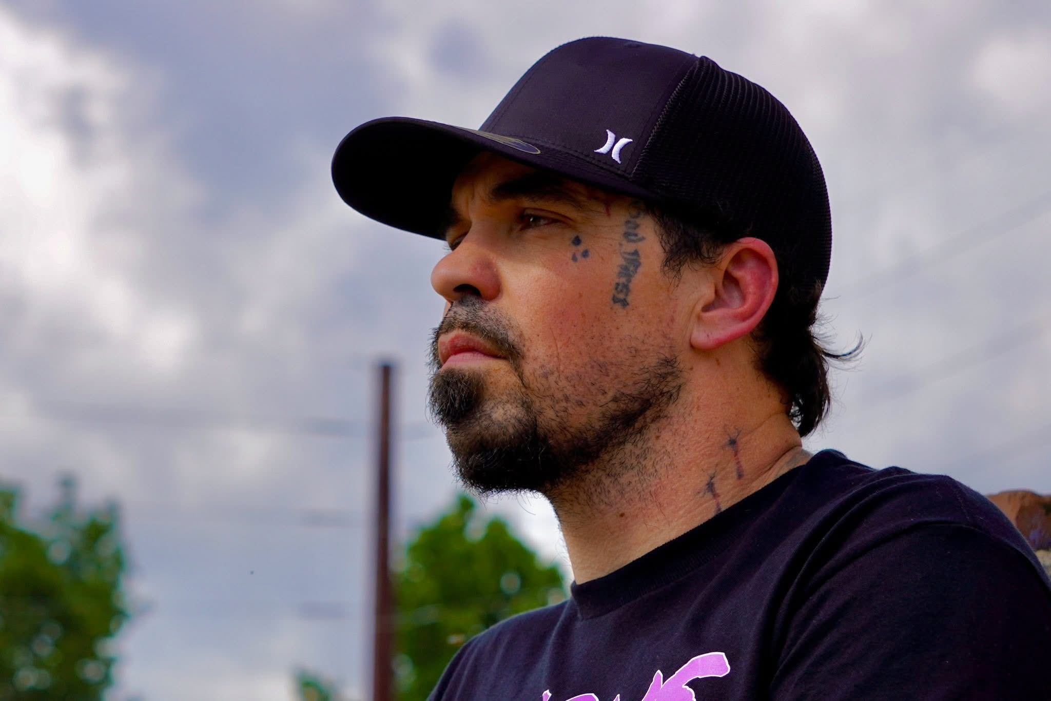 Man with a beard and tattoos wearing a black cap and black shirt, looking to the side outdoors with cloudy sky