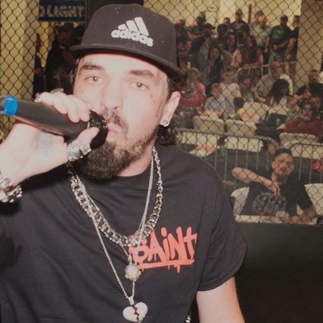 Man wearing a black cap and multiple necklaces, holding a microphone, with a crowd behind a chain-link fence.