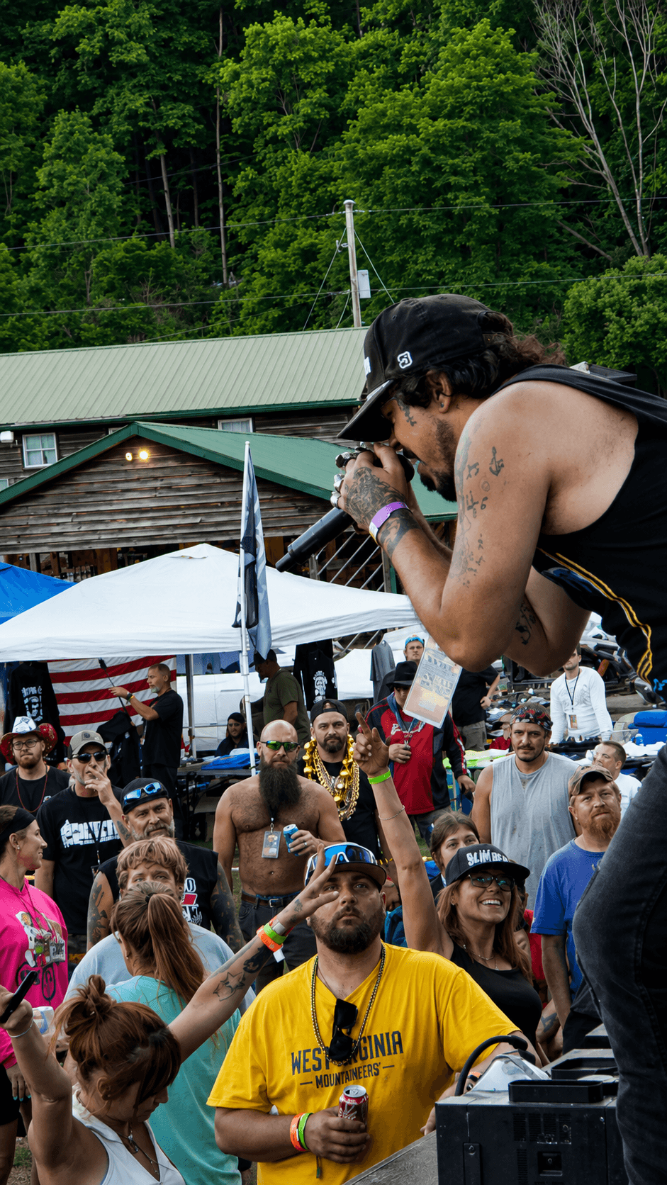 Performer with long hair, sunglasses, and tattoos sings into a microphone at an outdoor event, crowd watching below.