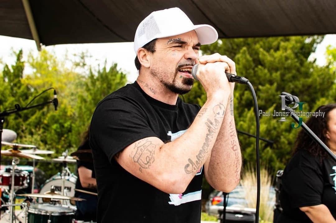 Man singing into microphone outdoors with trees and band equipment in background.