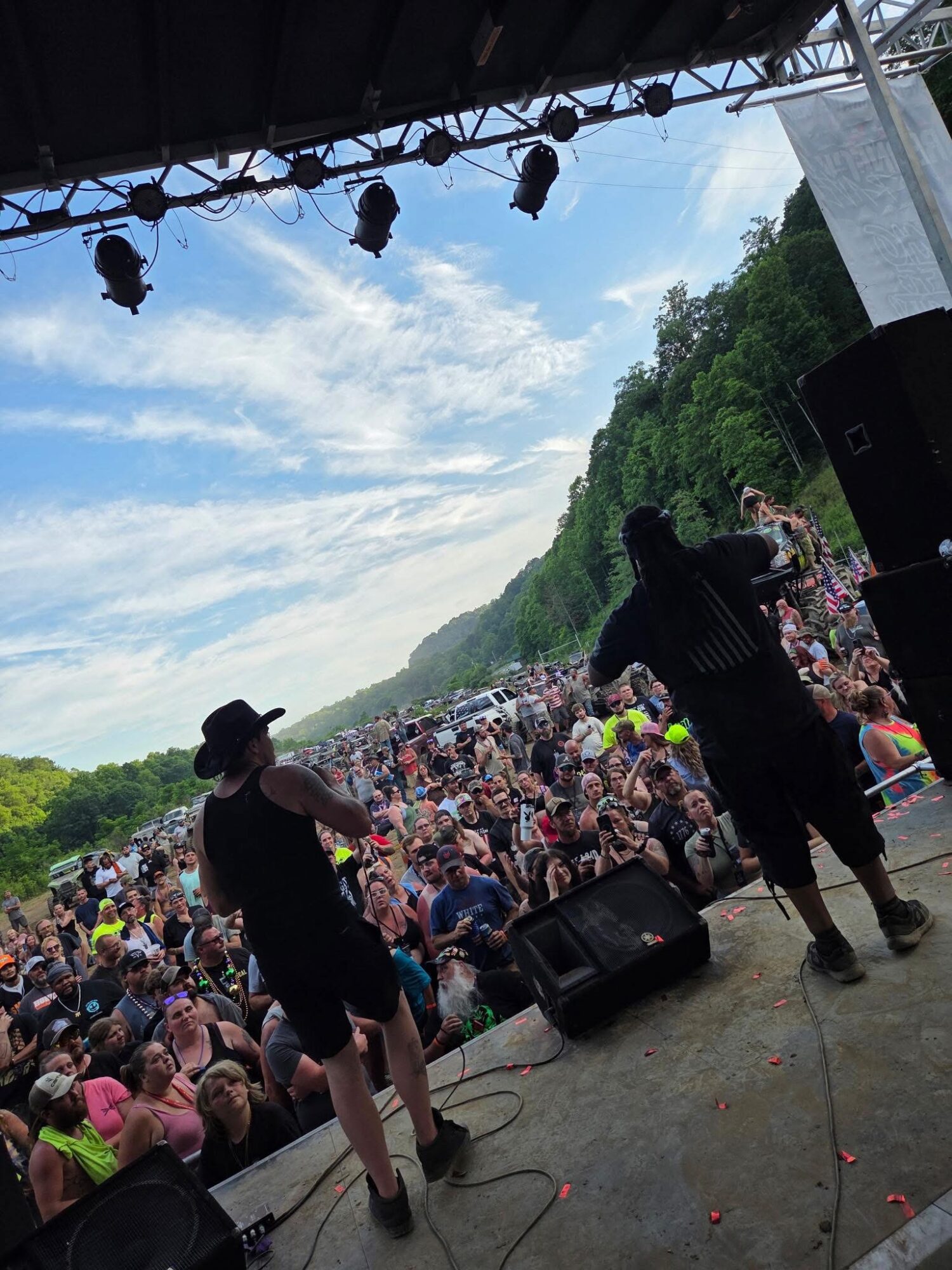 Performers on stage facing a large outdoor crowd with trees and blue sky in the background.