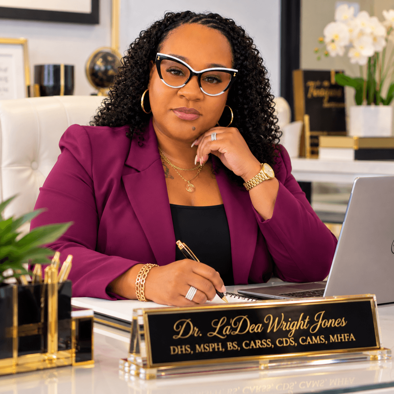 Woman with curly hair, glasses, and jewelry, sitting at desk with laptop and nameplate, in an office setting.