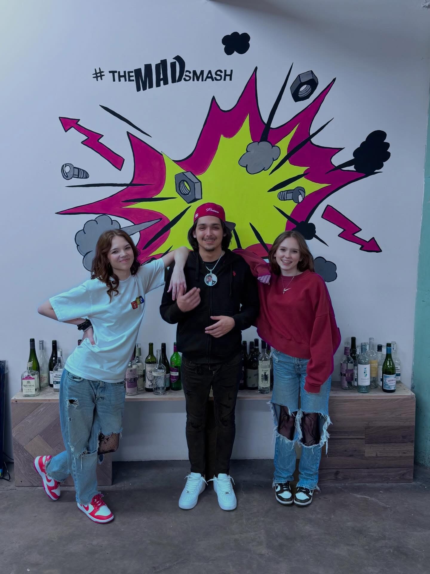 Three people stand in front of a colorful comic-style wall with bottles on a shelf below. Two girls and a woman smile.
