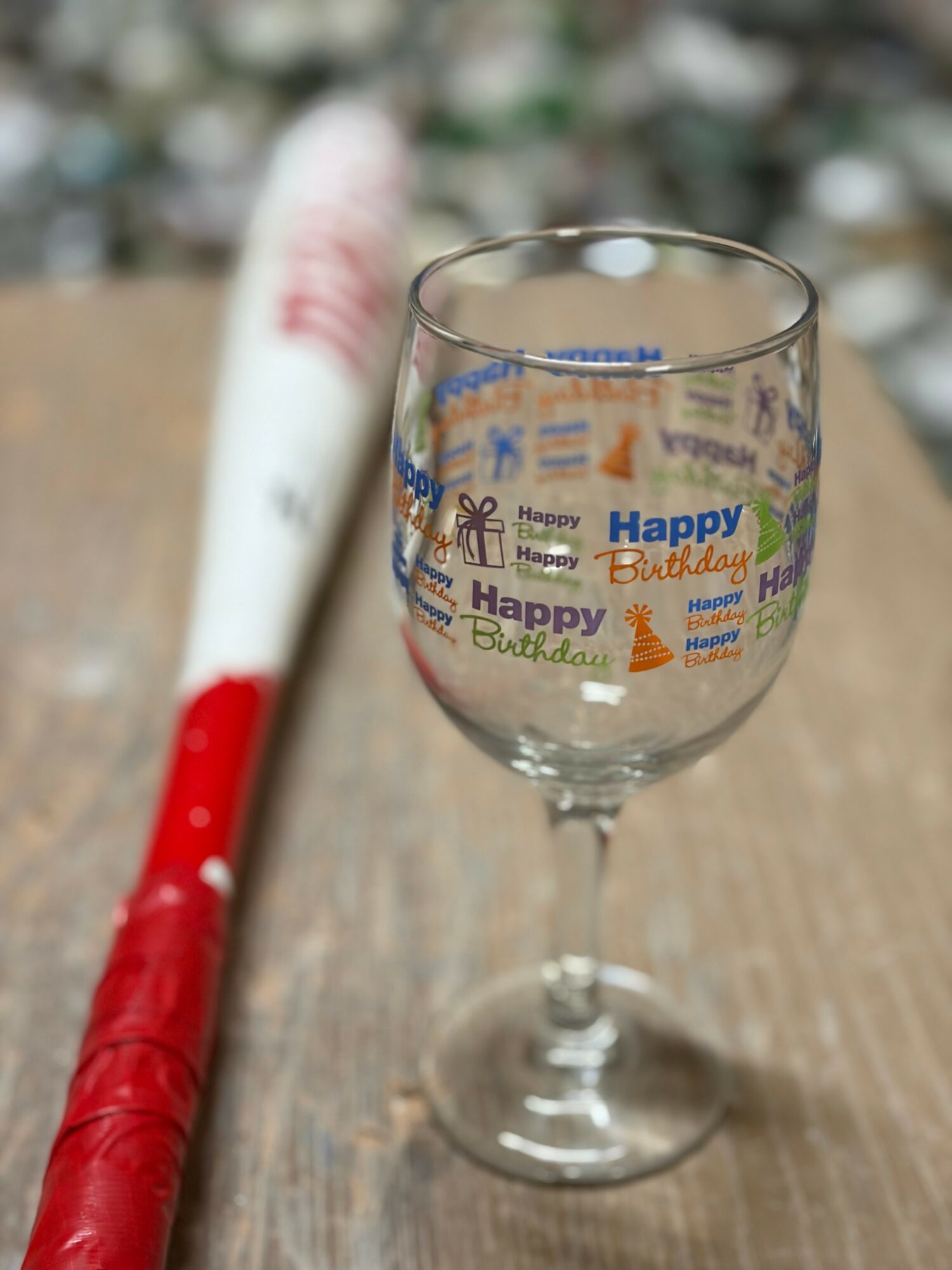 A wine glass with birthday-themed stickers on a wooden surface, with a red and white object in the background.
