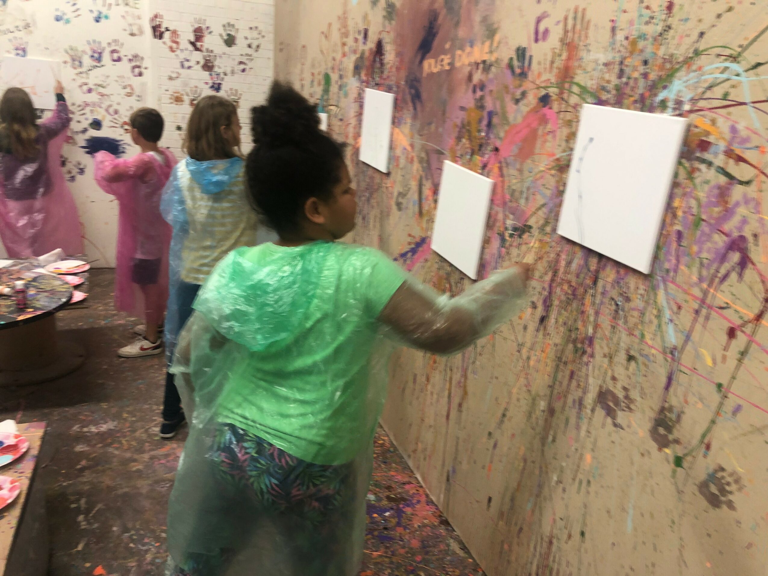 Children painting on a large wall with colorful splashes and four blank canvases, some children wearing smocks, in an art room.