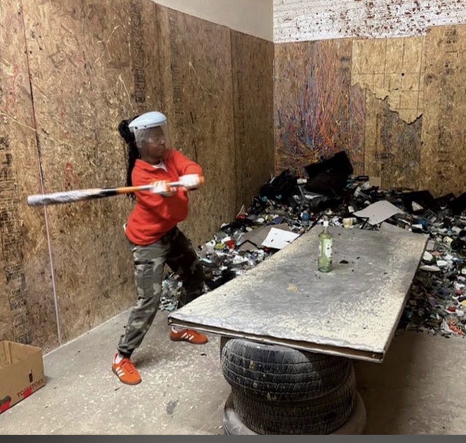 Child in red shirt and cap holding a stick, standing in a cluttered room with a table and debris.