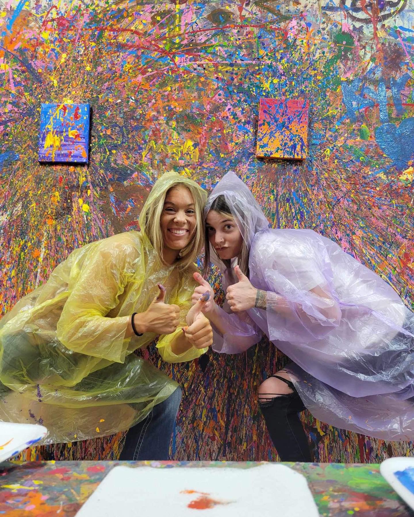 Two women in rain ponchos giving thumbs up in front of colorful abstract wall art.