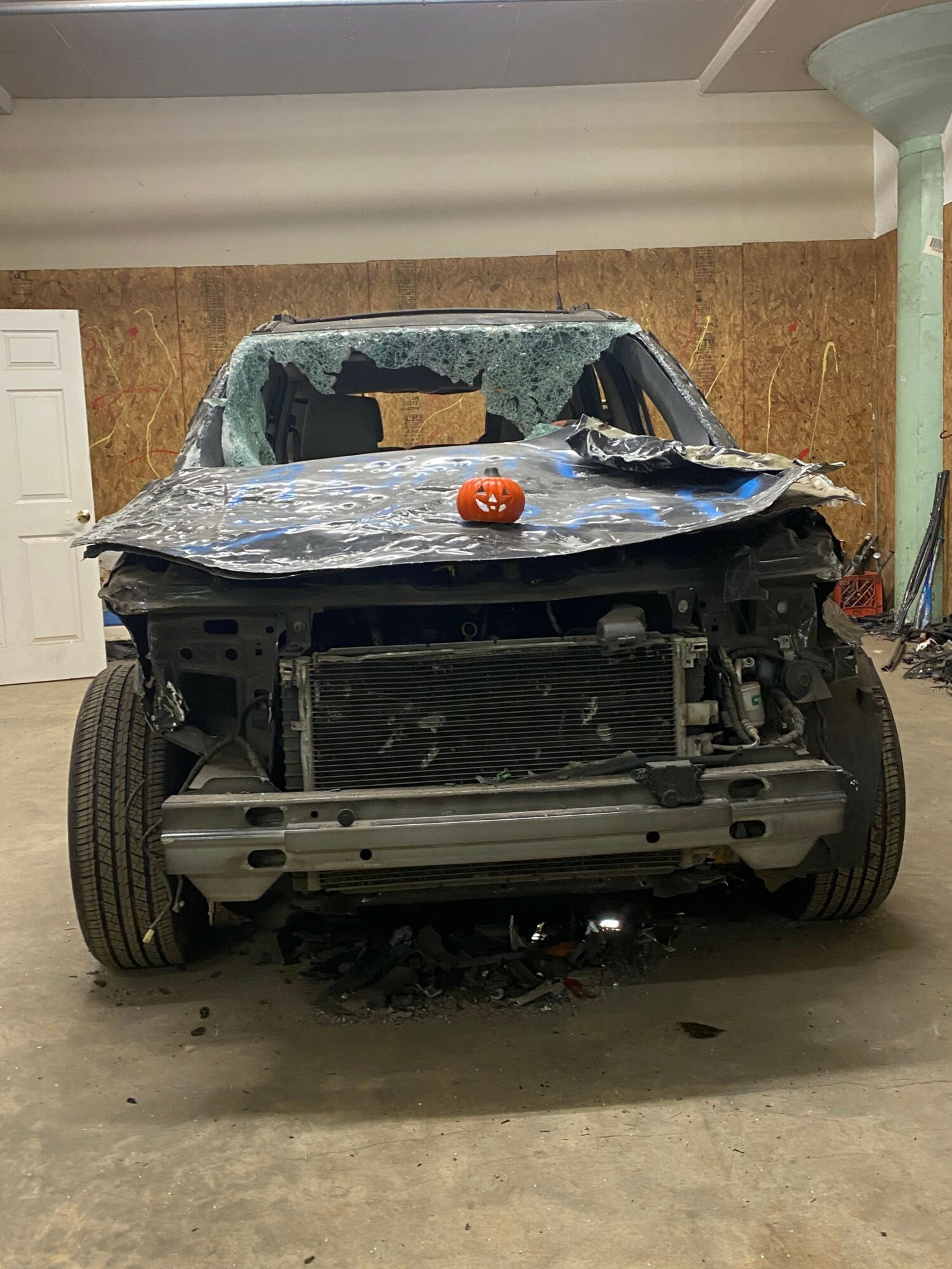 A vehicle with a damaged front, parked in a garage with a concrete floor and wooden wall, covered with a tarp.