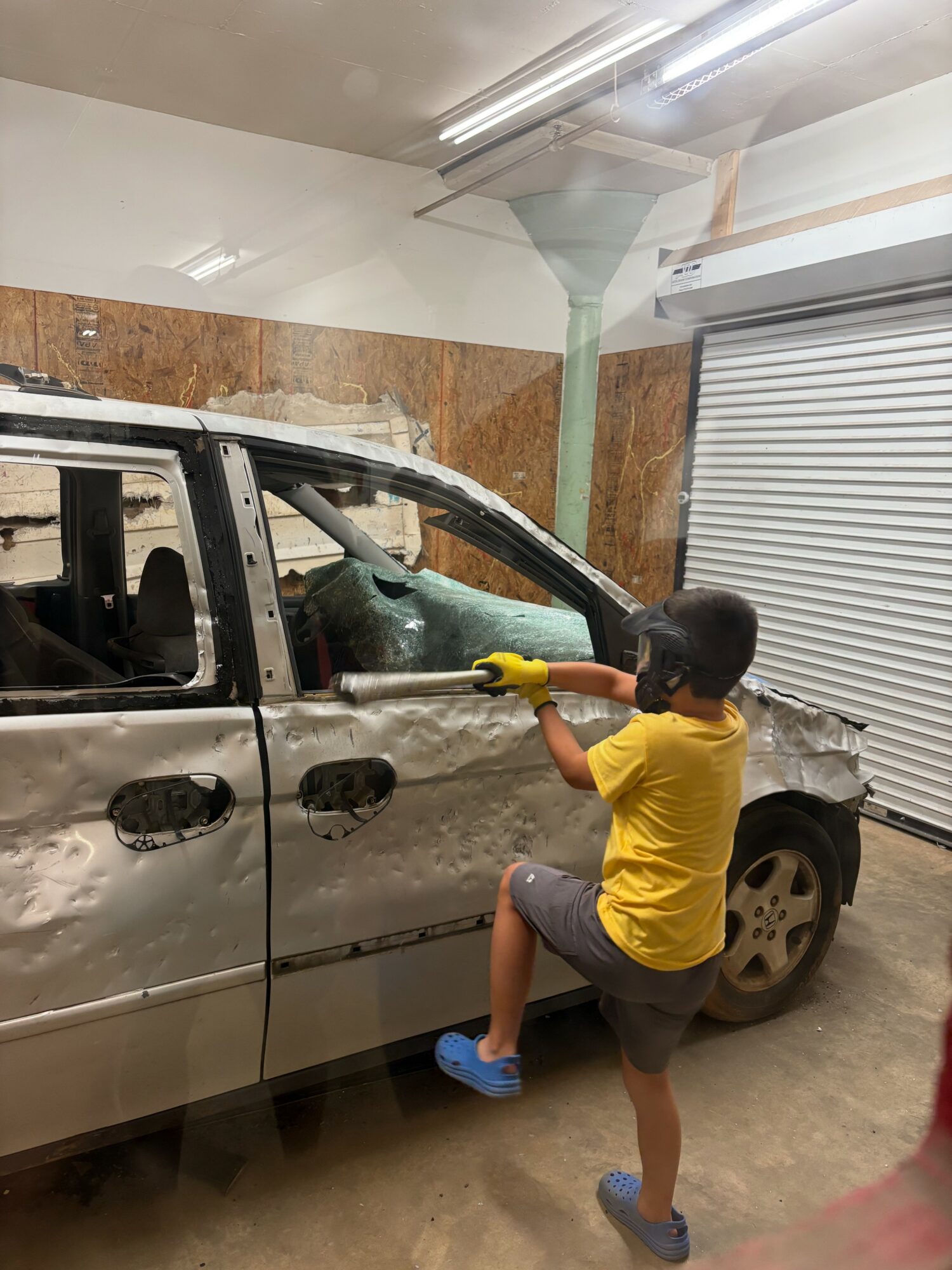 Child in yellow shirt and shorts using a power tool on a damaged silver car inside a garage.
