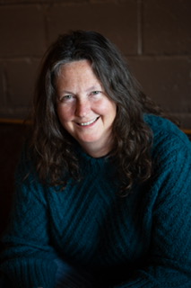 Woman with long dark hair smiling, wearing a dark teal sweater, against a dark background.