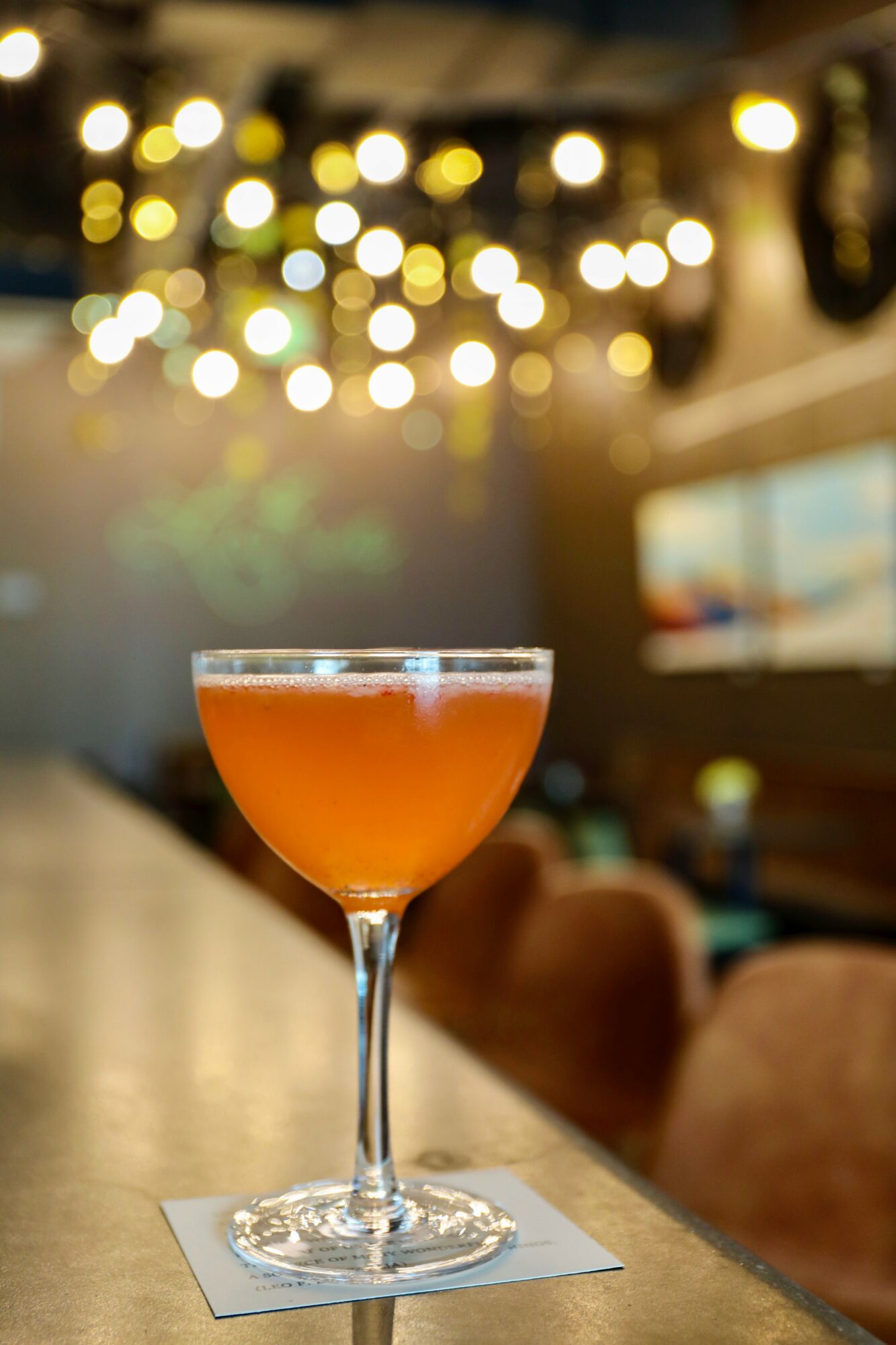 A cocktail glass with an orange drink on a bar counter, blurred warm lights in background.