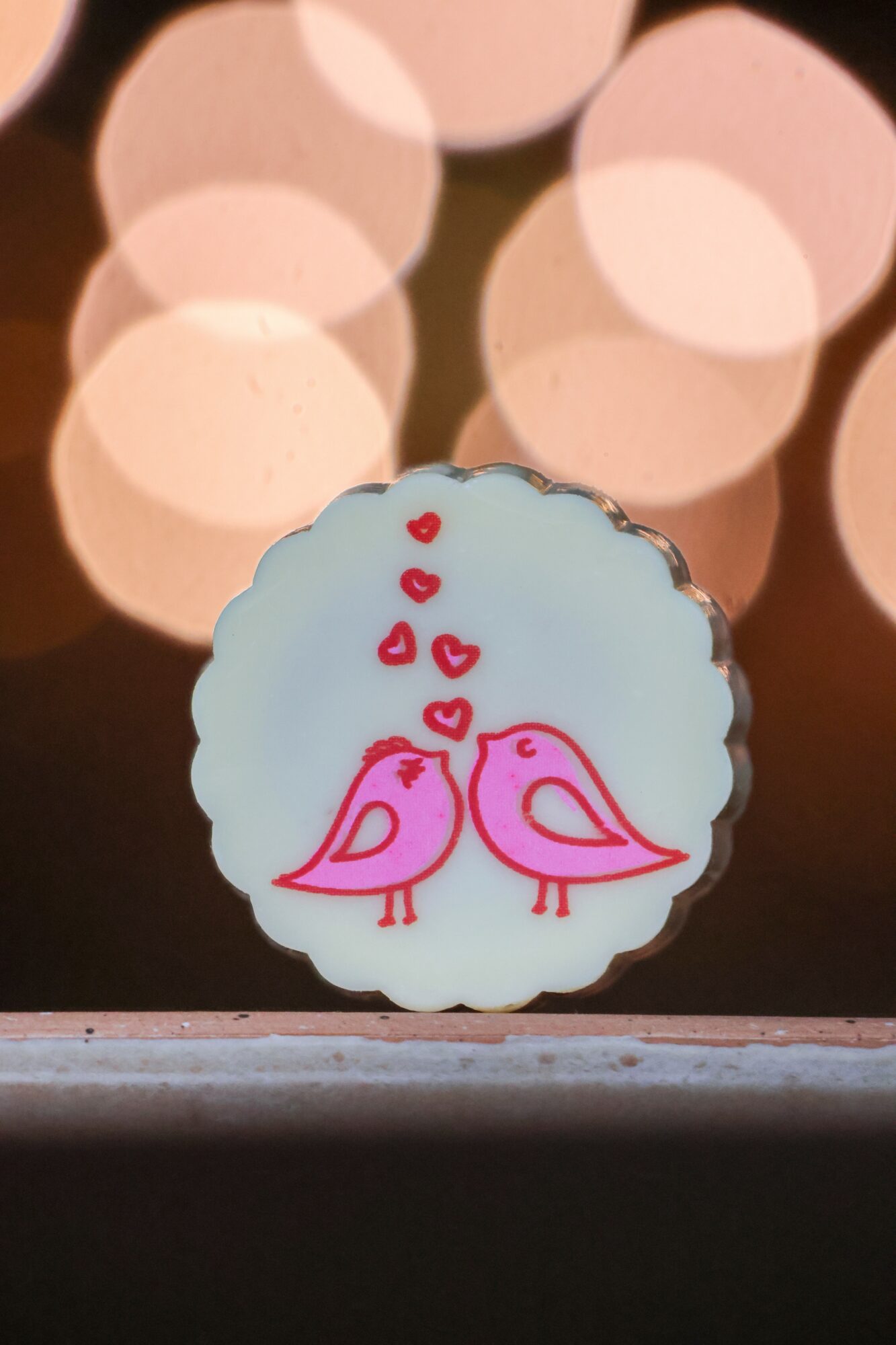 A round cookie with a white background features two pink birds facing each other with hearts above them, against a blurred background of lights.
