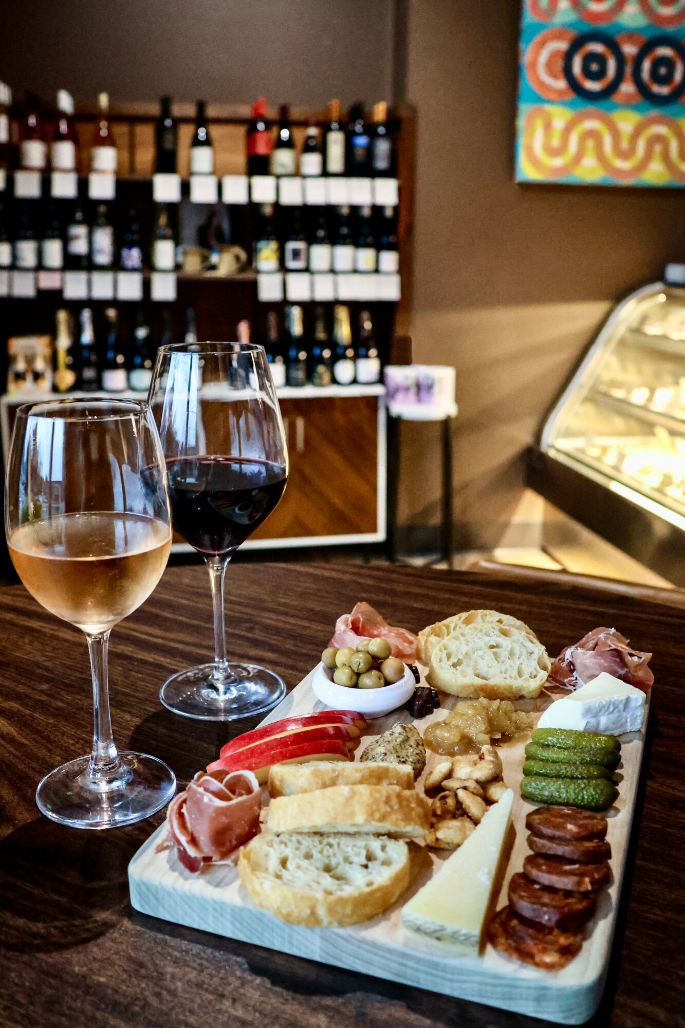 Wine glasses and a cheese platter on a table in a wine shop, with shelves of wine bottles in the background.