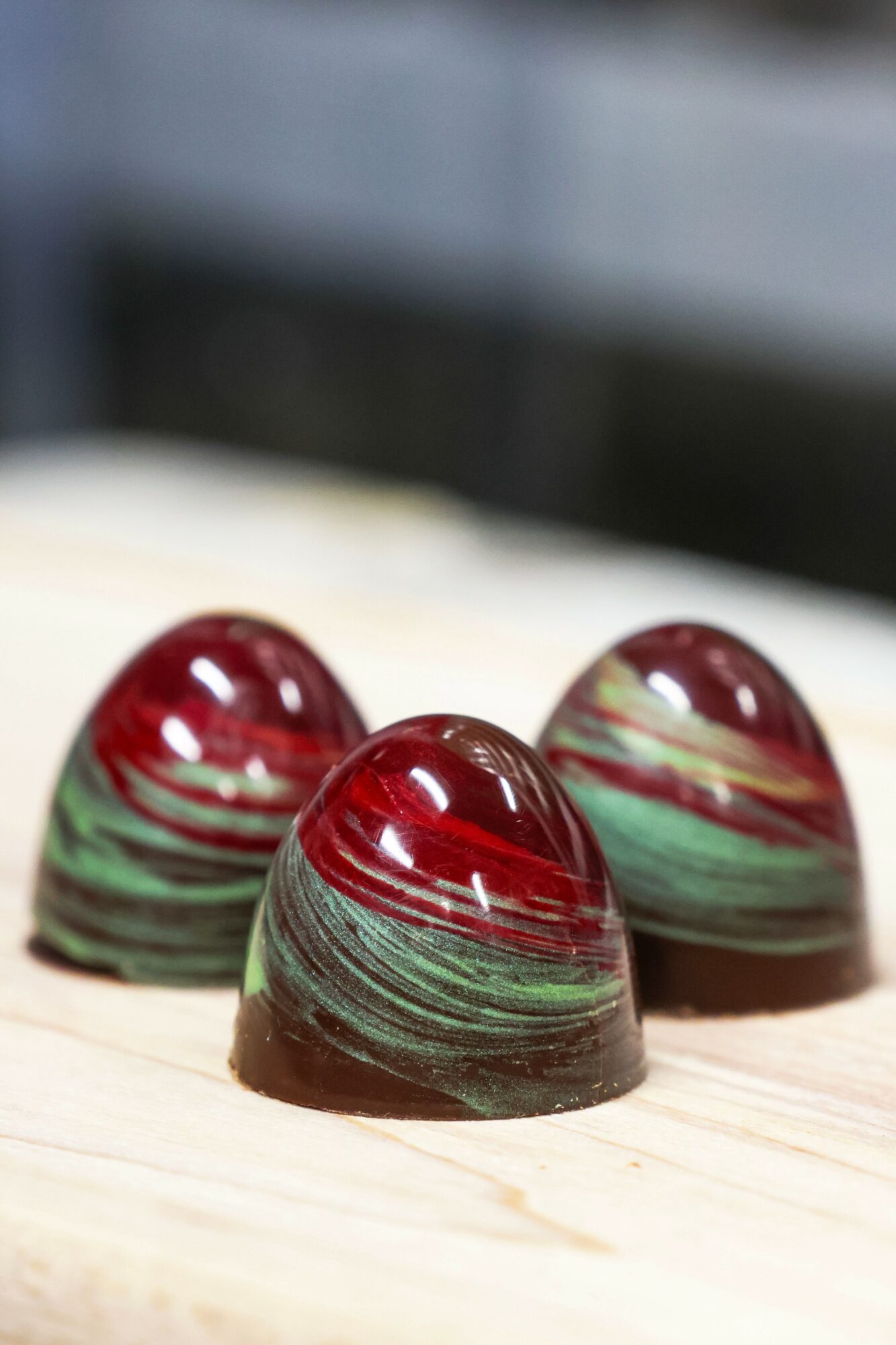 Three dome-shaped chocolates with red and green swirled patterns on a light wooden surface.