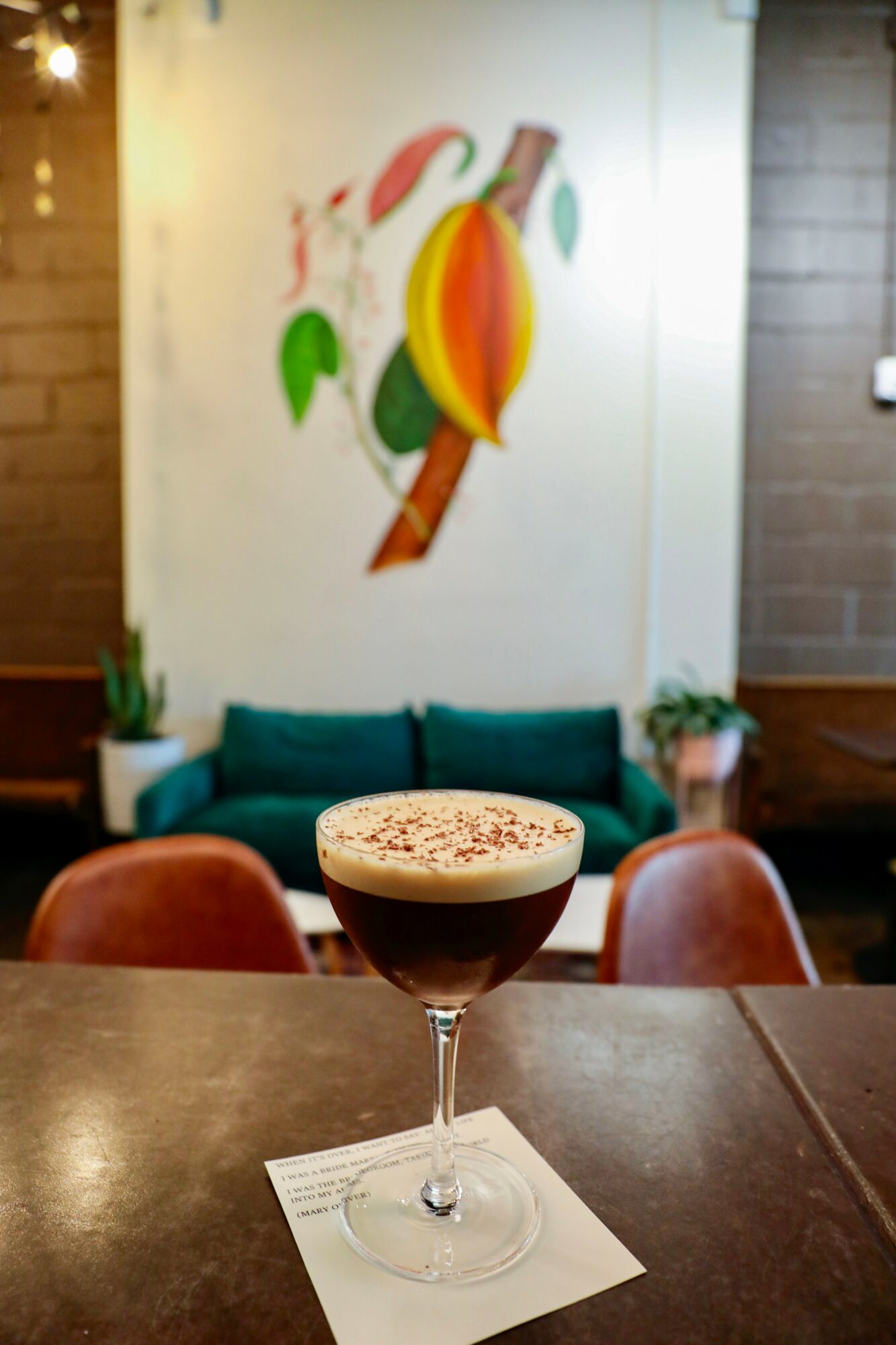 A layered coffee drink in a glass with foam and chocolate sprinkles, on a napkin on a table, with a colorful abstract painting behind.