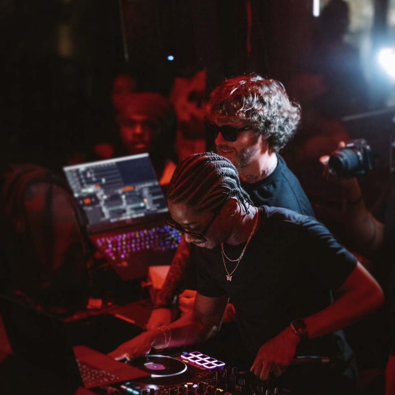 Two people working with DJ equipment in a dimly lit setting, with a laptop and a mixer visible.