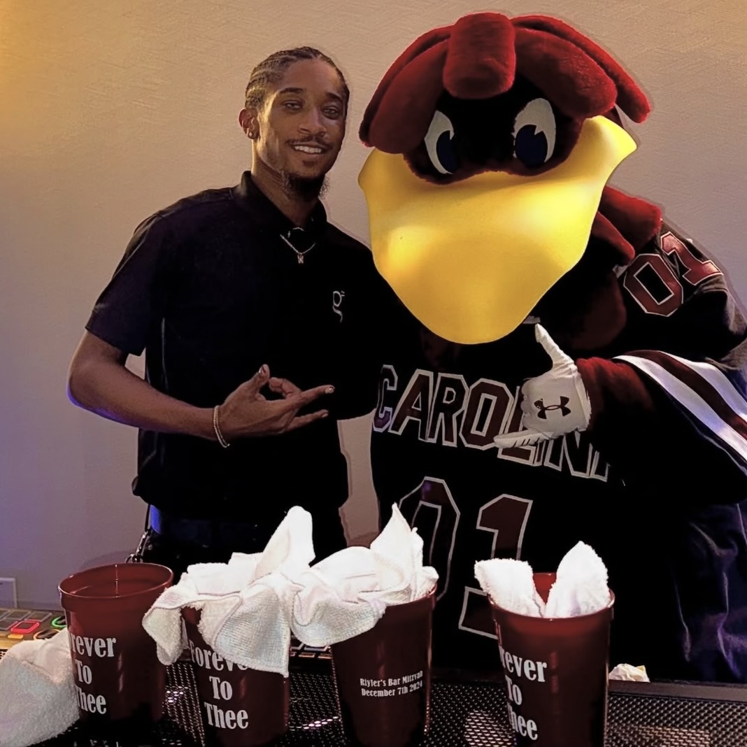 A person standing next to a large mascot with a yellow beak and red hair, holding cups with napkins, in an indoor setting.