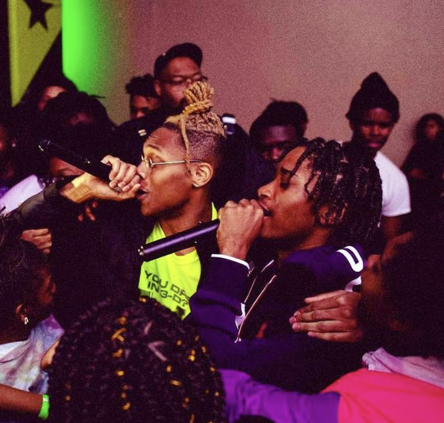 Two young people with microphones perform on stage, surrounded by a group of people, some with glasses and braided hair.