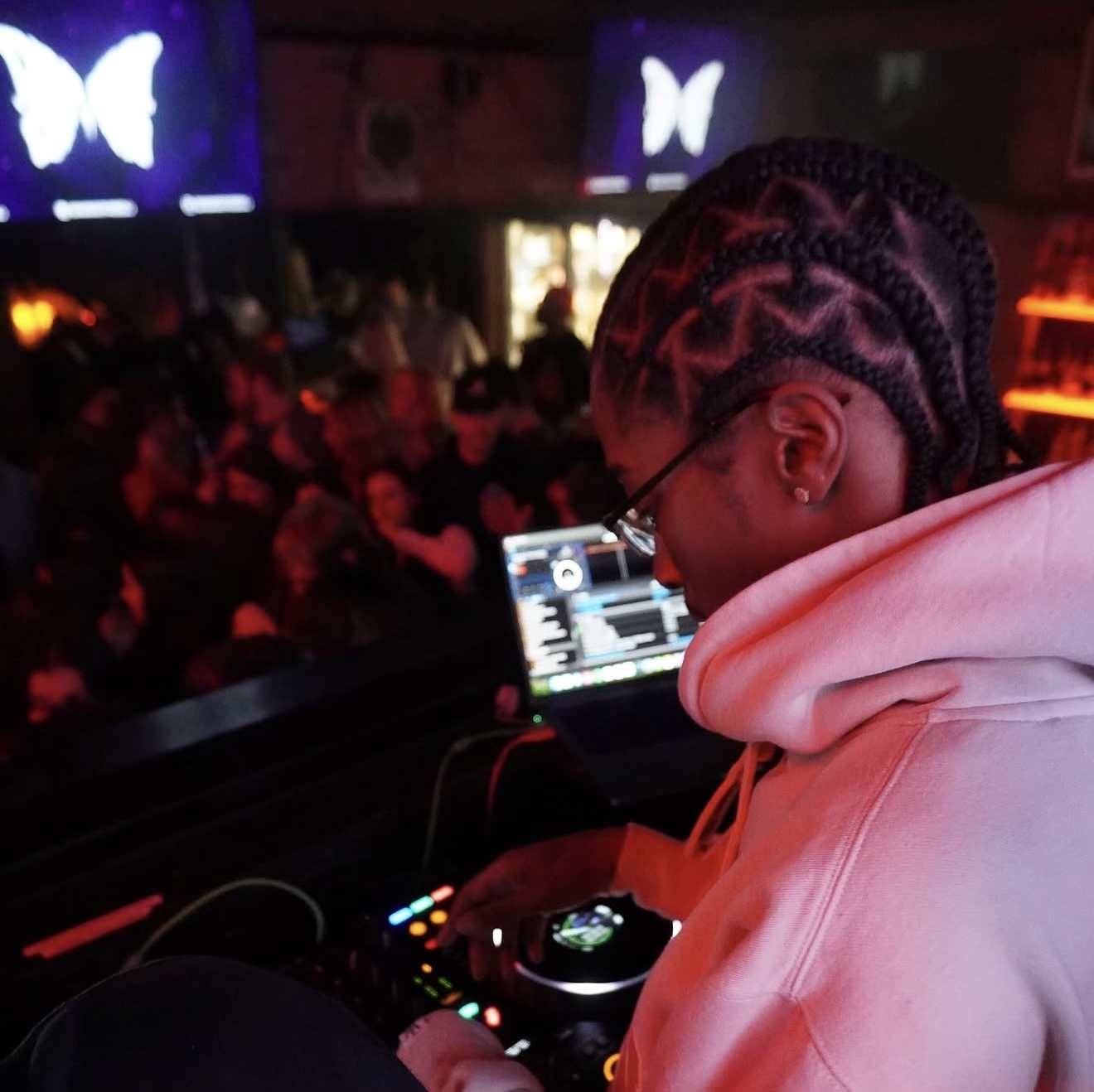 DJ with braided hair and glasses performs at a nightclub with a crowd and butterfly screens in background.